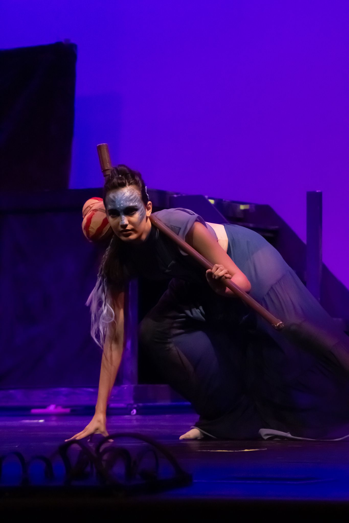 A solo dancer in dramatic face paint crouches on stage holding a staff under purple lighting.