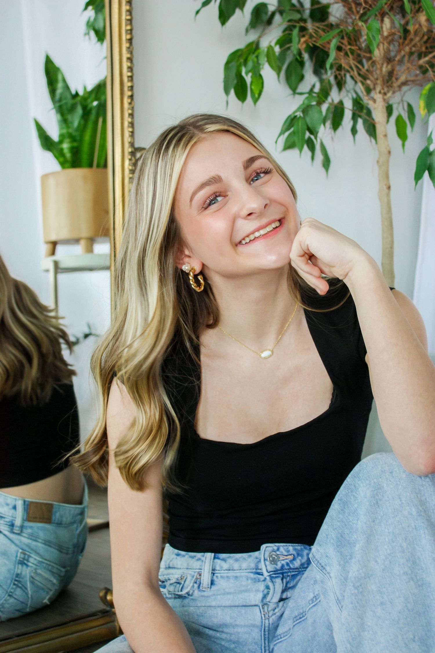 Smiling blonde woman in black tank top and jeans, wearing gold earrings, seated near a mirror and green plant.