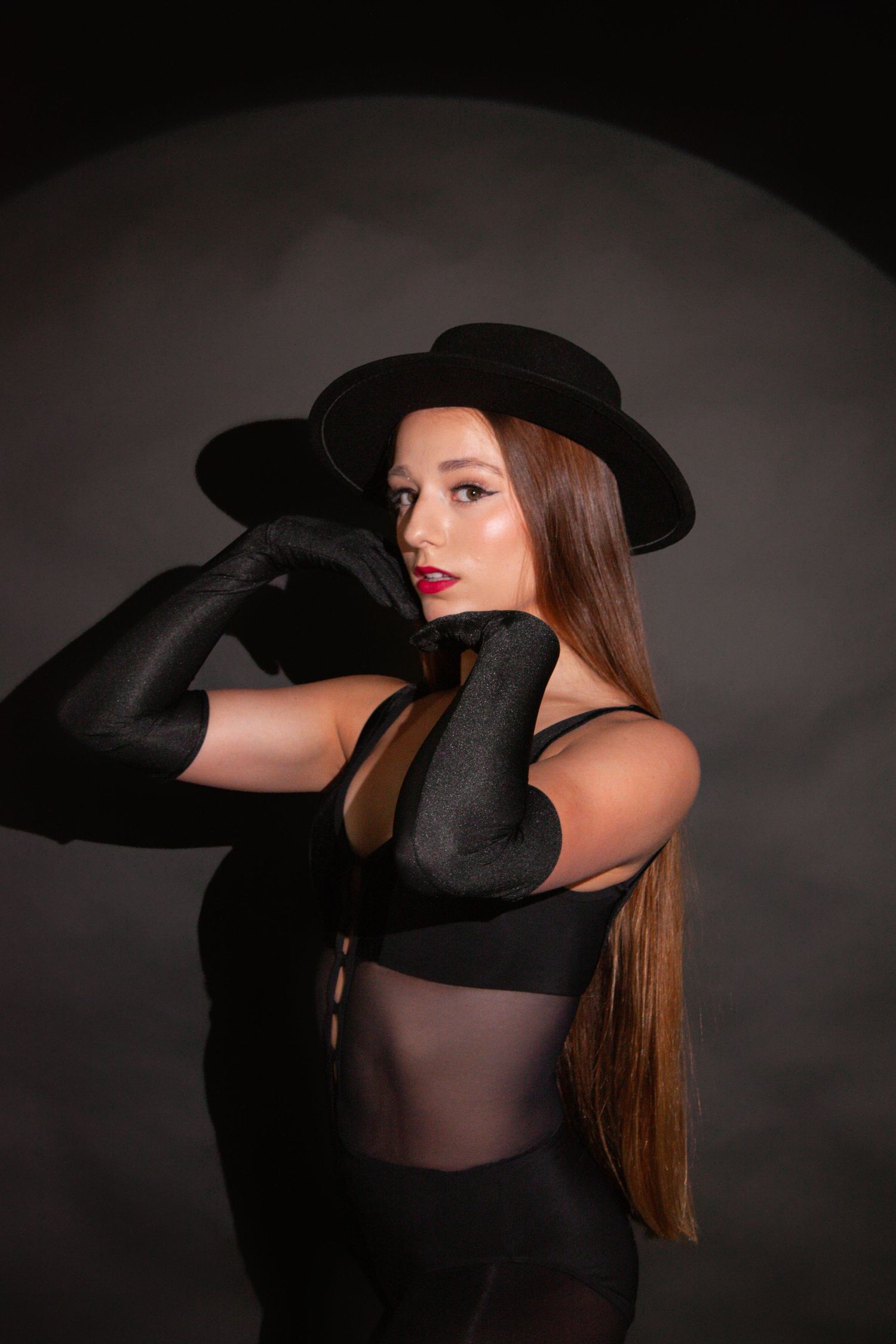 Woman in black hat and gloves poses elegantly in dark dramatic studio portrait setting.