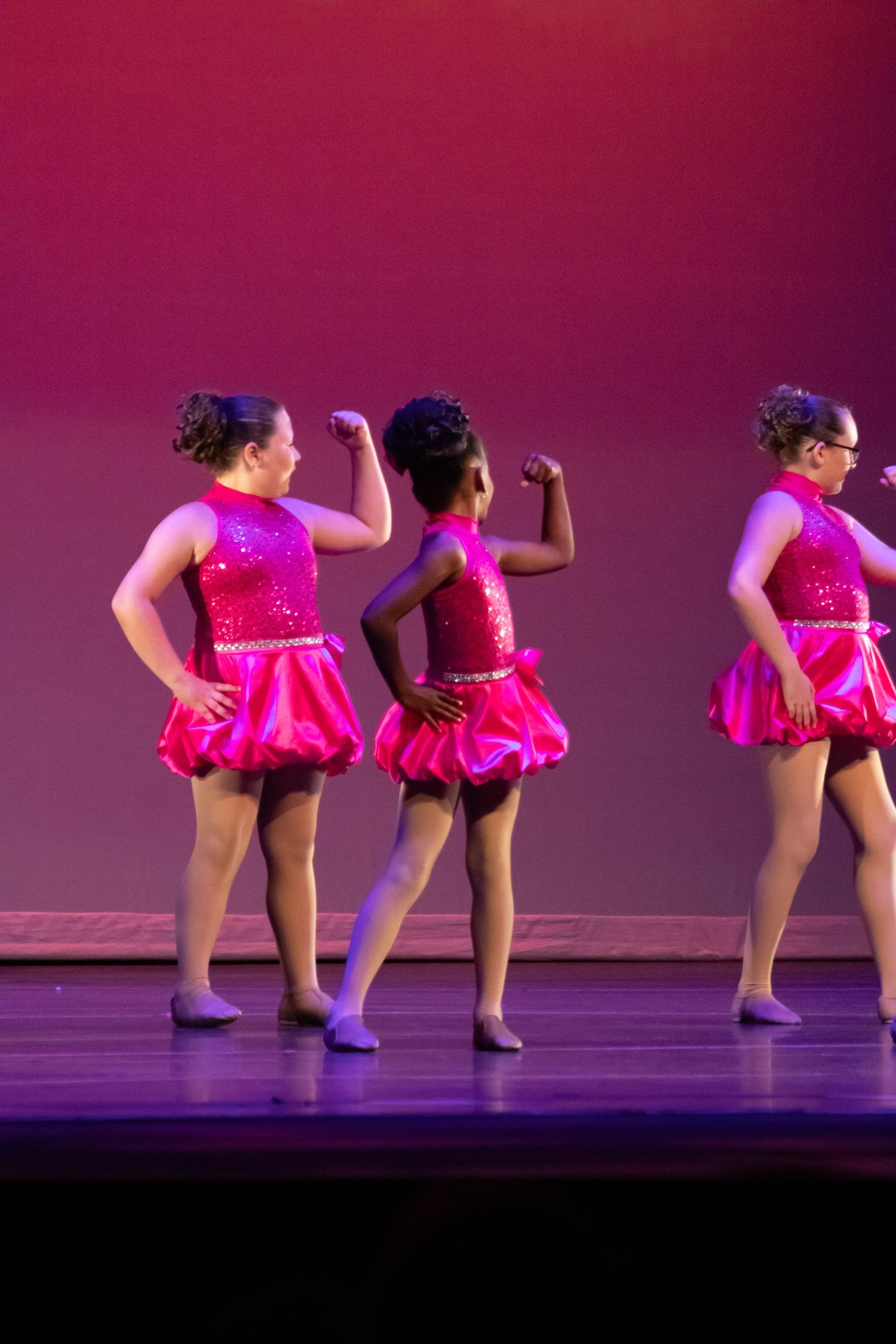 Three girls in pink sequin costumes flex their muscles on stage during a dance recital performance.