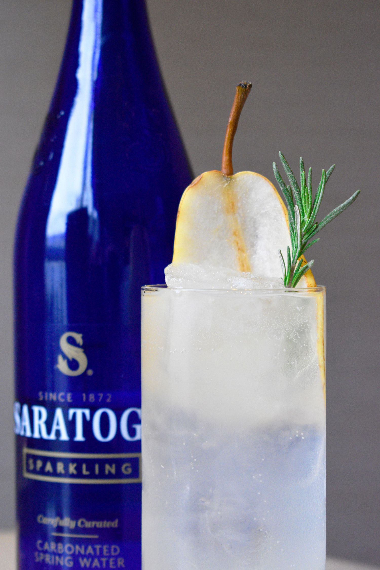 Saratoga sparkling water highball cocktail garnished with a fresh pear slice and rosemary sprig over crushed ice.