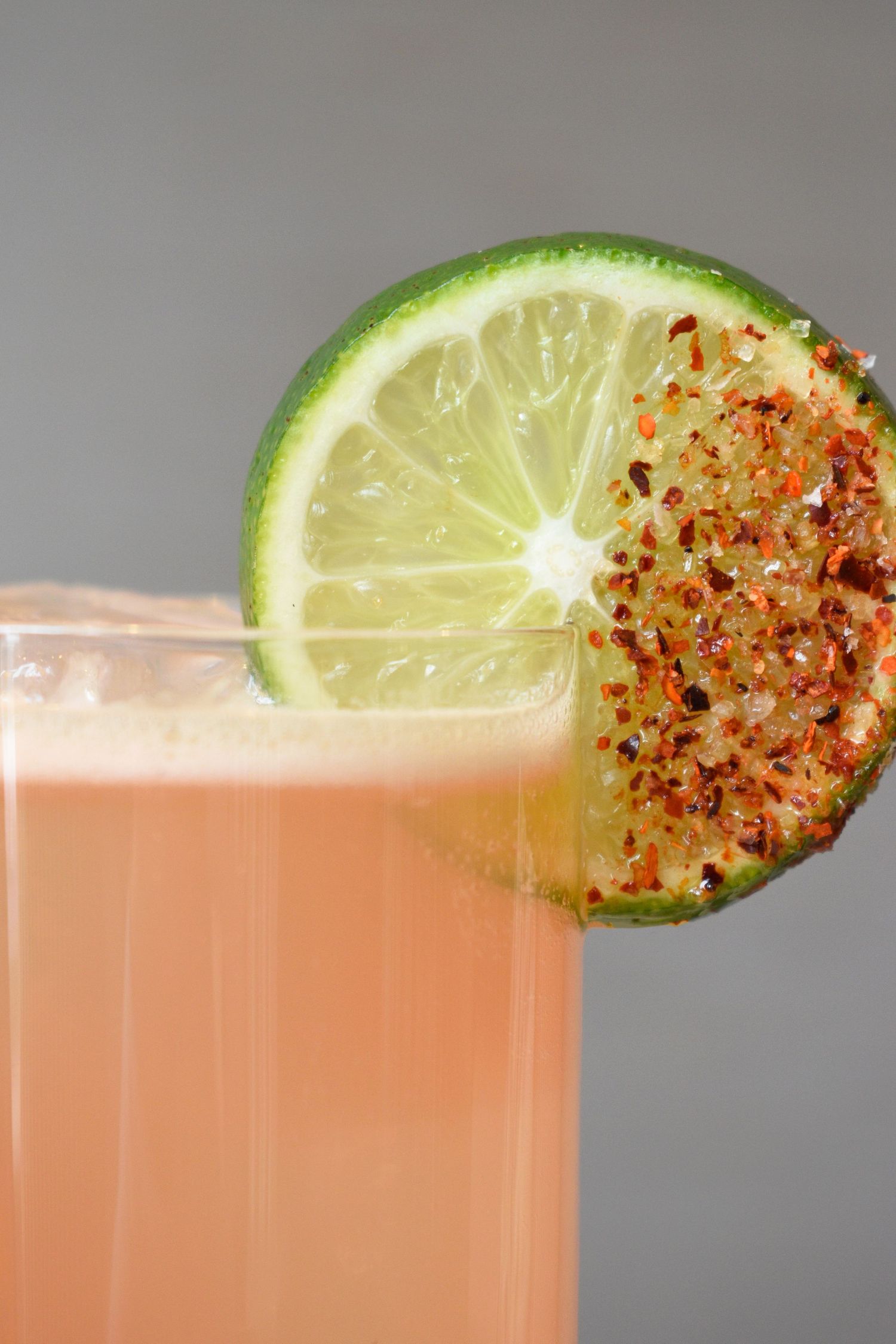 A pink cocktail garnished with a lime slice coated in red chili flakes on the rim of the glass.