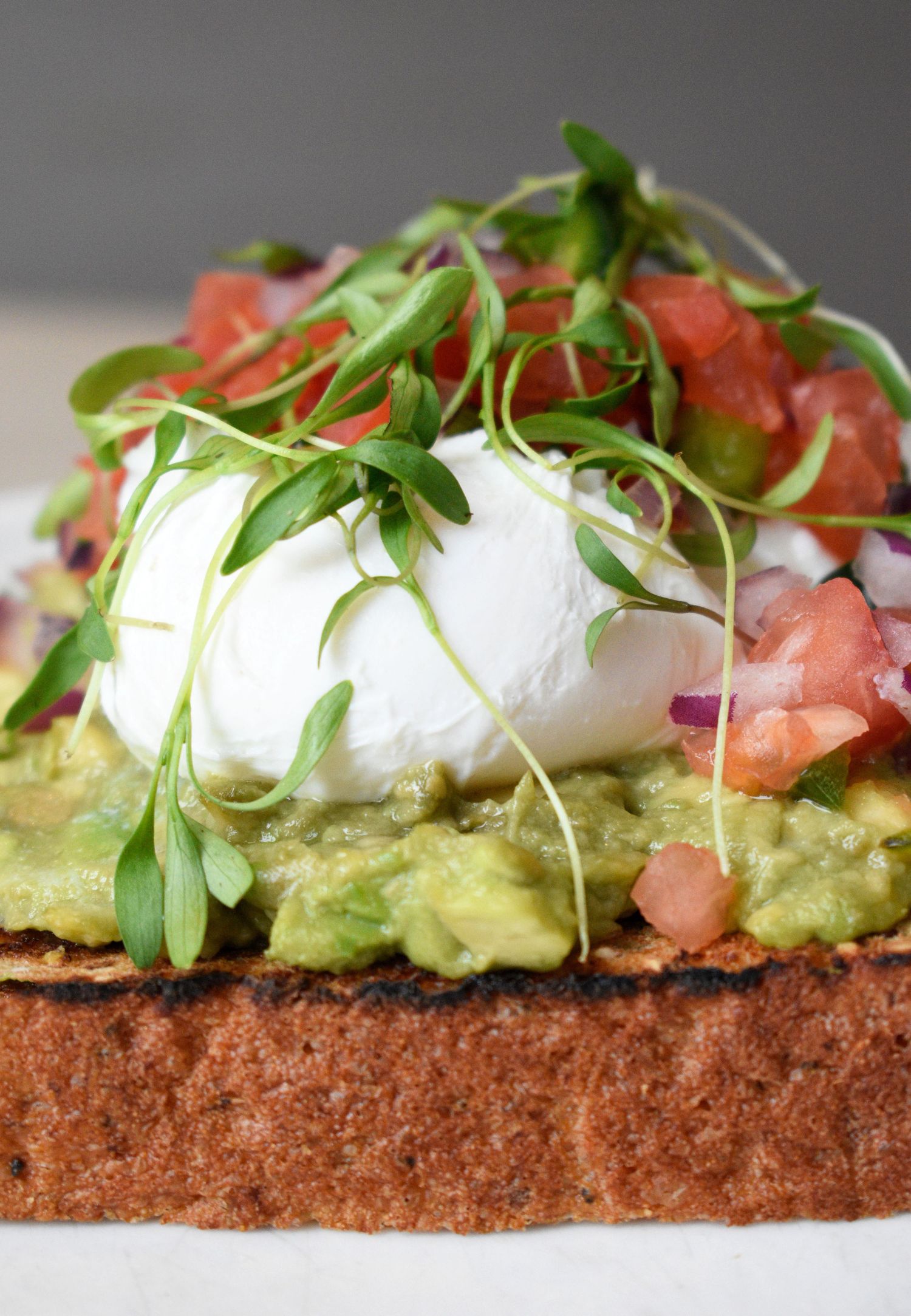 Close-up of avocado toast topped with a poached egg, fresh pico de gallo, and microgreens on toasted bread.