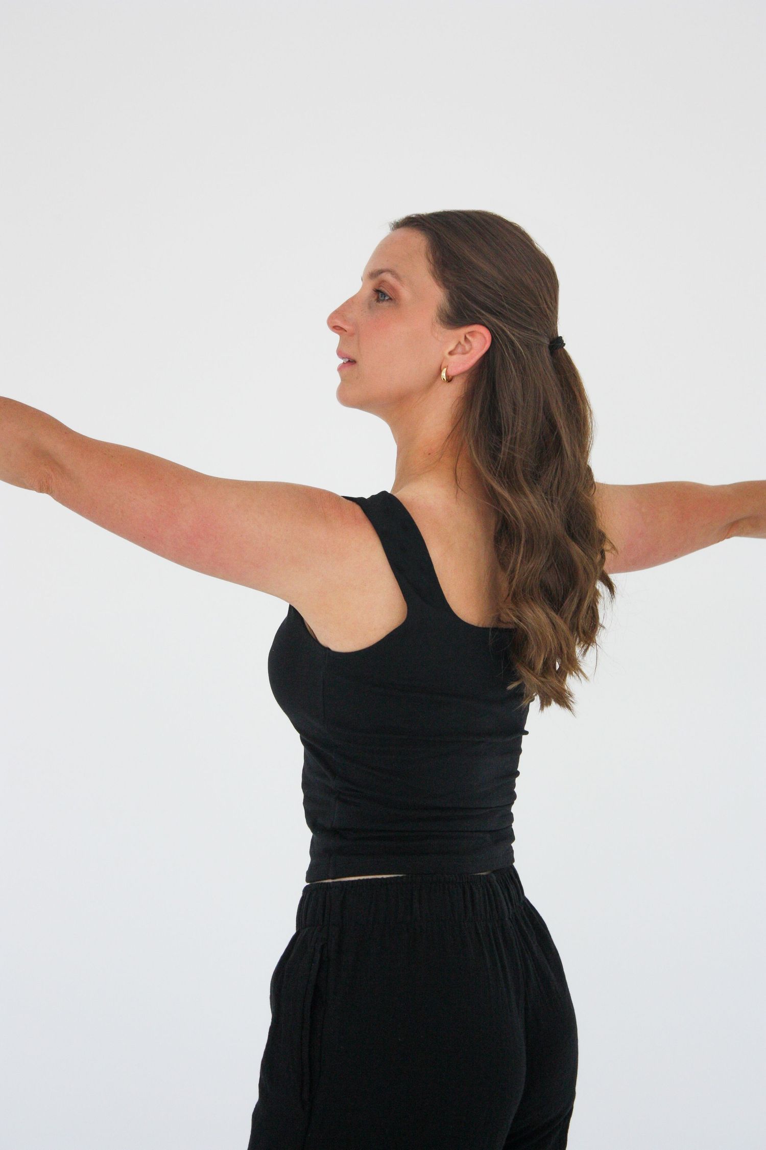 Woman in black tank top and pants stands with arms outstretched in a graceful dance pose against white background.