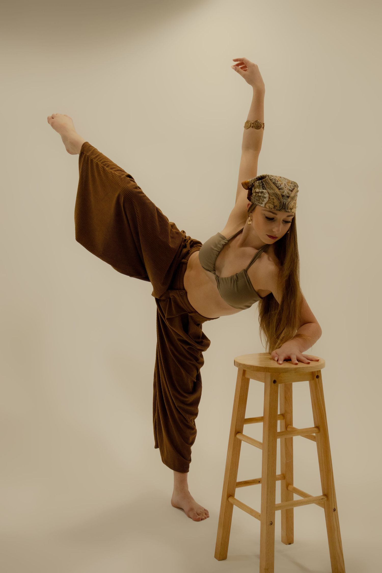 Flexible dancer in brown harem pants and crop top performs high kick pose beside a wooden stool.
