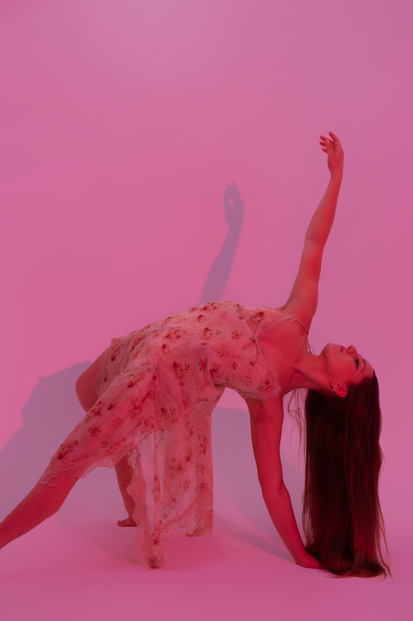 Dancer in floral dress doing side stretch pose in pink-lit studio.