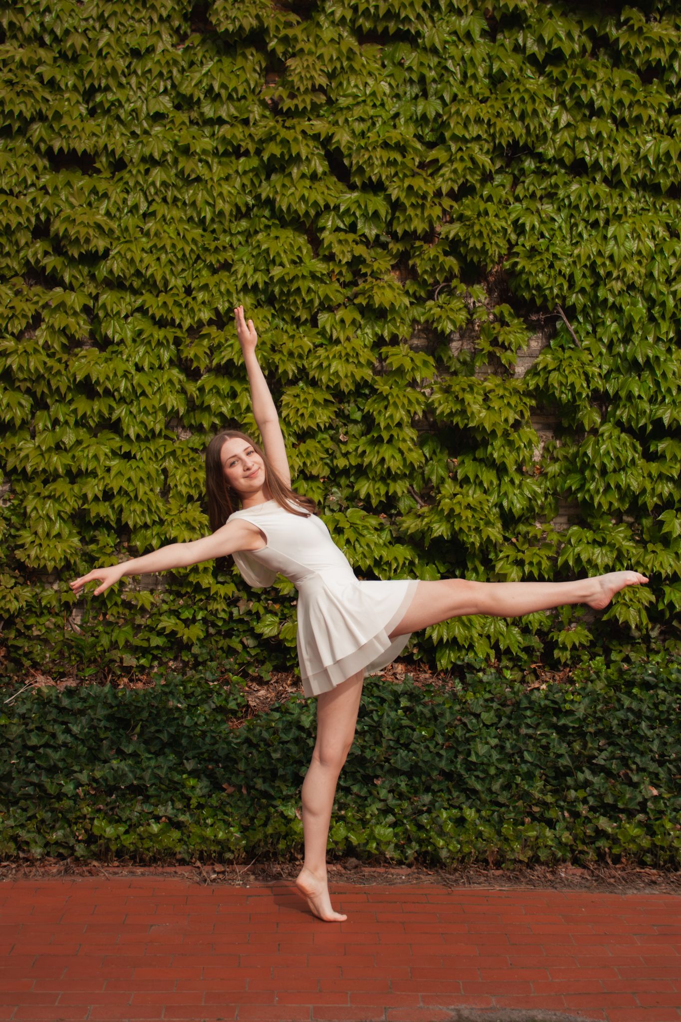 A graceful ballerina in a white dress performs an arabesque pose against a lush green ivy wall.