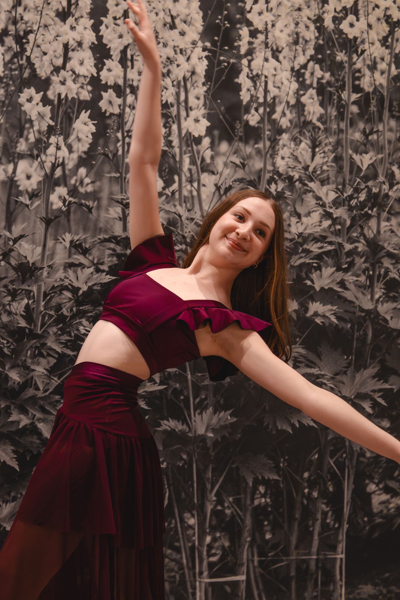 A smiling dancer in a burgundy crop top and skirt performs an expressive pose against a floral backdrop.
