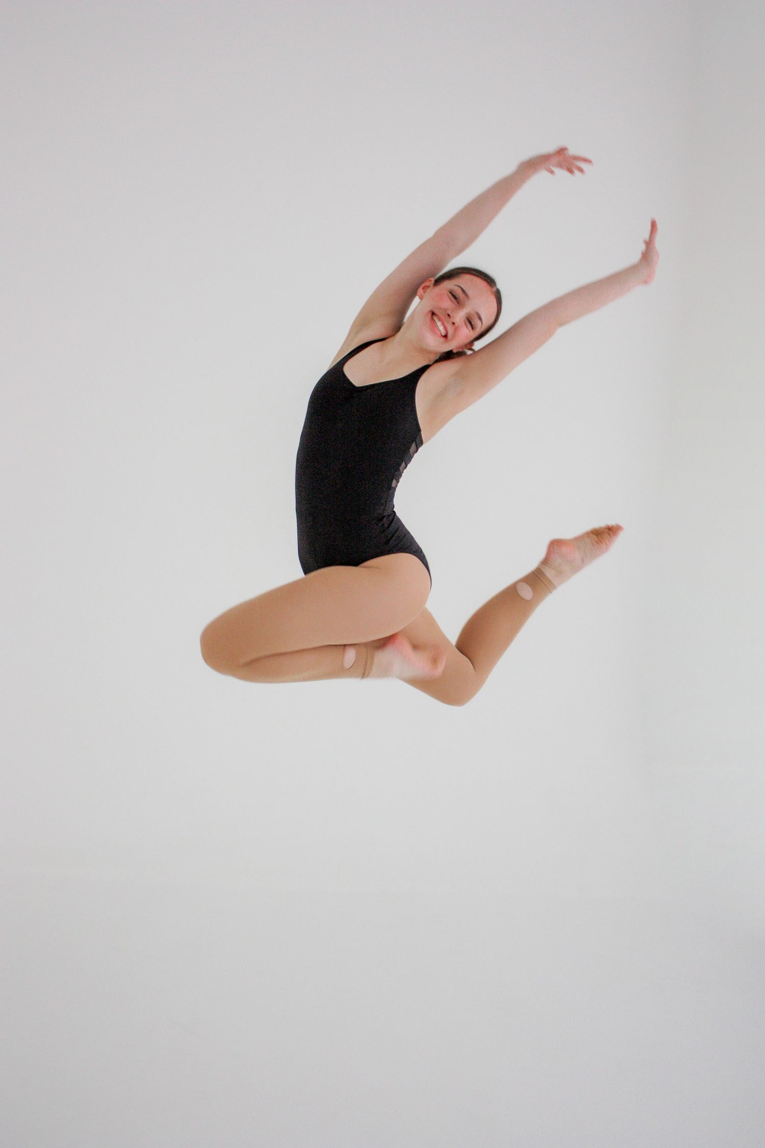 A smiling female dancer in a black leotard performs a joyful leap jump against a white studio background.
