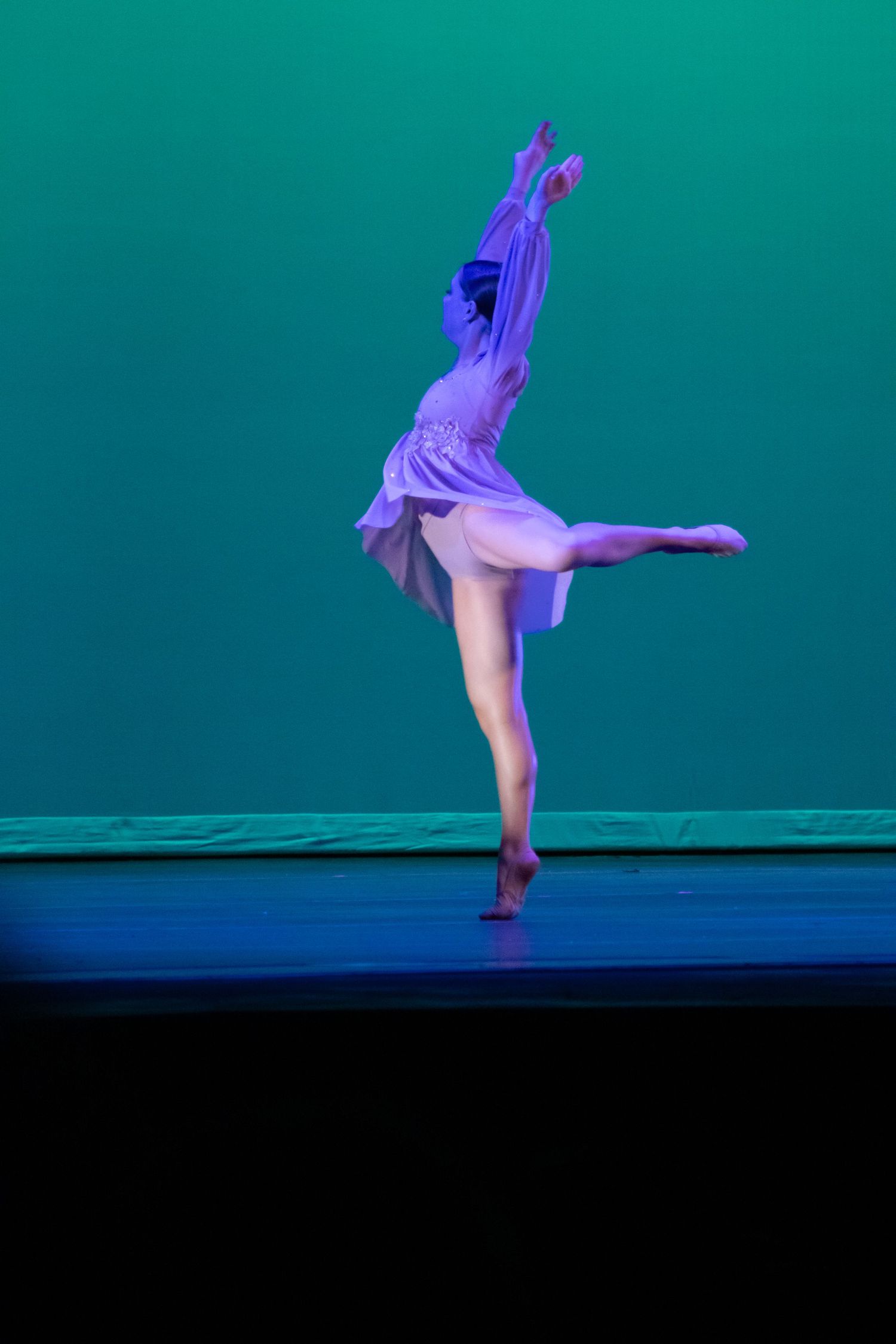 A ballet dancer performs an arabesque on stage in a white dress under teal and blue stage lighting.