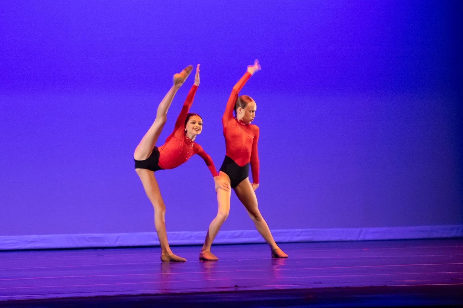 Two dancers in red leotards and black shorts perform synchronized moves on a blue-lit stage.