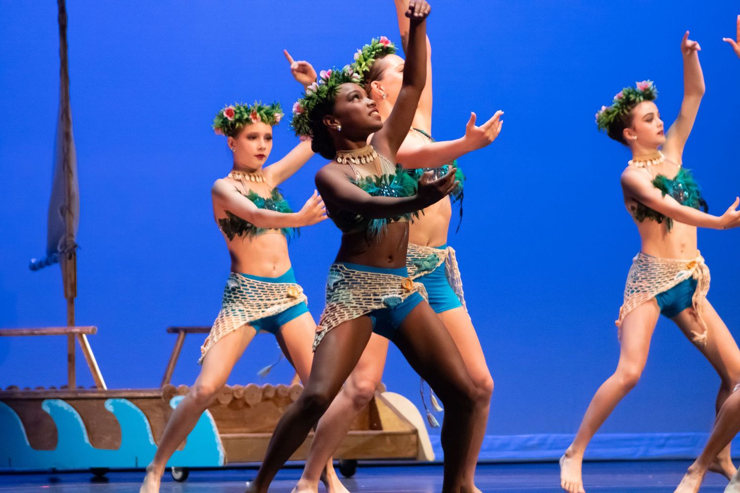 Three dancers in teal costumes perform an energetic routine on a blue-lit stage.