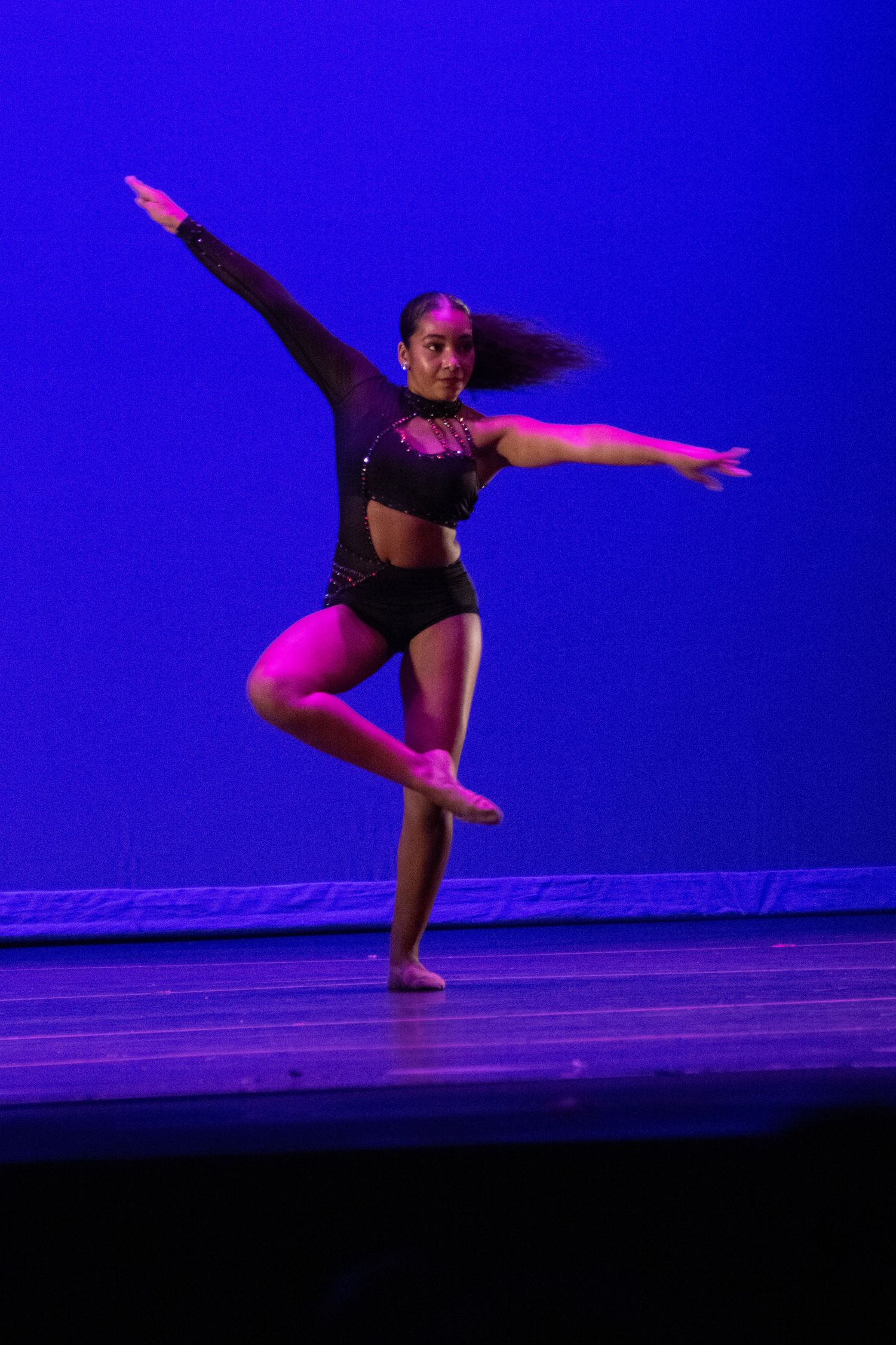 A dancer in black performs on stage under pink and blue lights, balancing on one leg with arms extended.