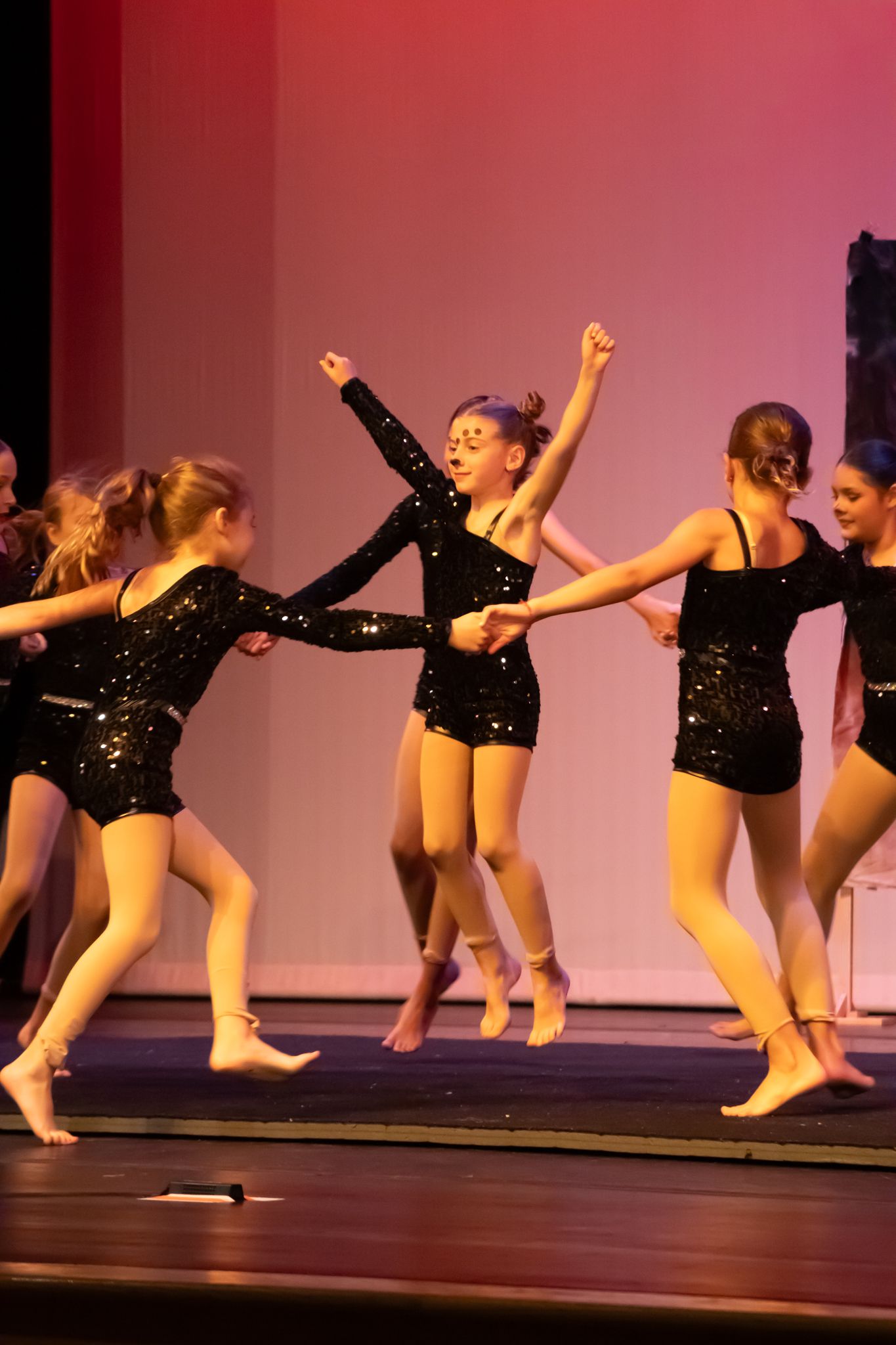 Girls in black sequined costumes dancing energetically on a red-lit recital stage.
