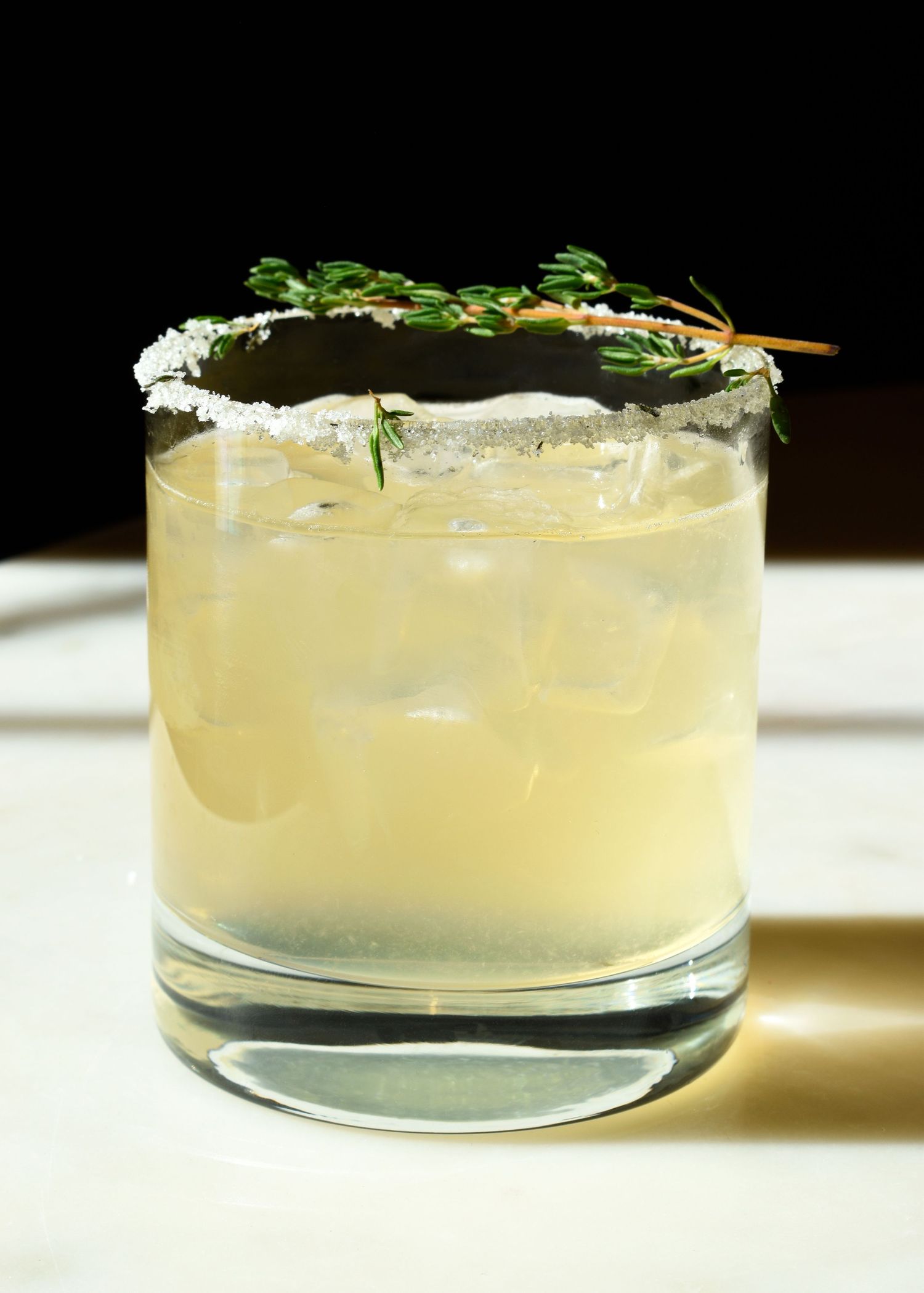 A salted rim rocks glass filled with pale yellow cocktail, ice cubes, and garnished with fresh thyme sprigs.