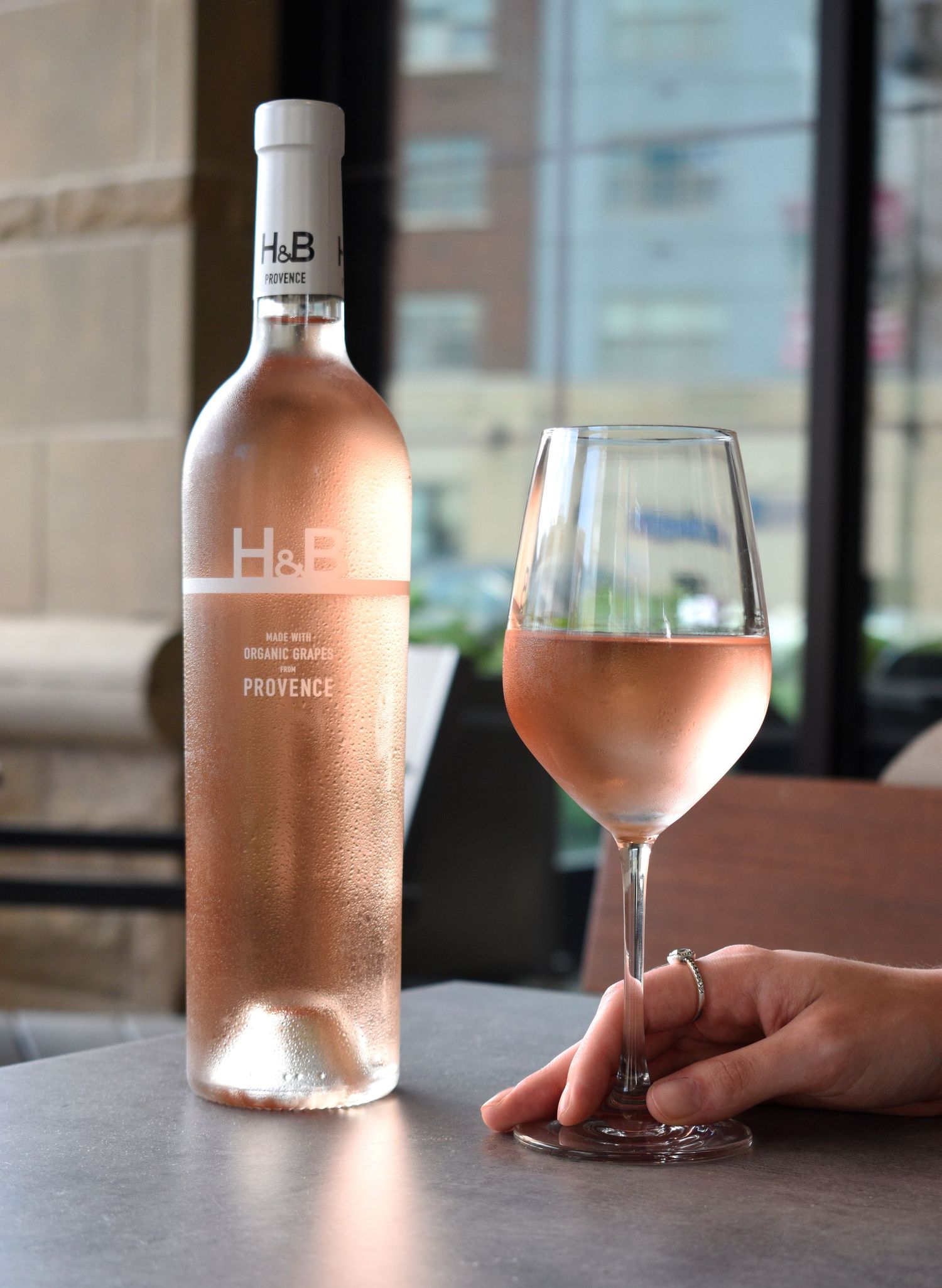 Chilled bottle of H&B Provence rose wine beside a glass of pink rose wine held by a hand.
