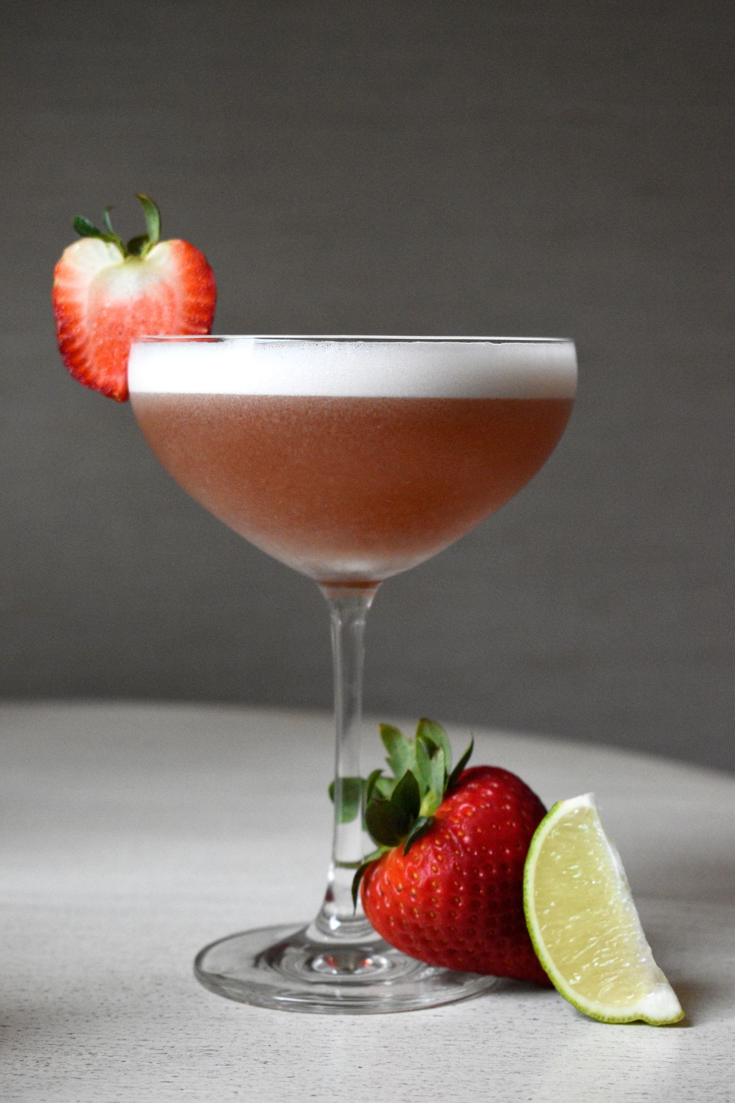 A frothy pink strawberry daiquiri in a coupe glass, garnished with a fresh strawberry and lime wedge.