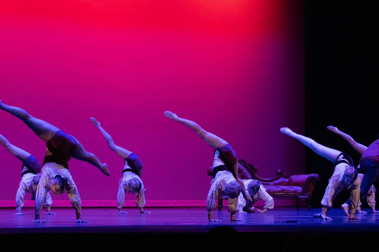 A line of dancers perform synchronized handstands with legs extended on a colorfully lit stage.