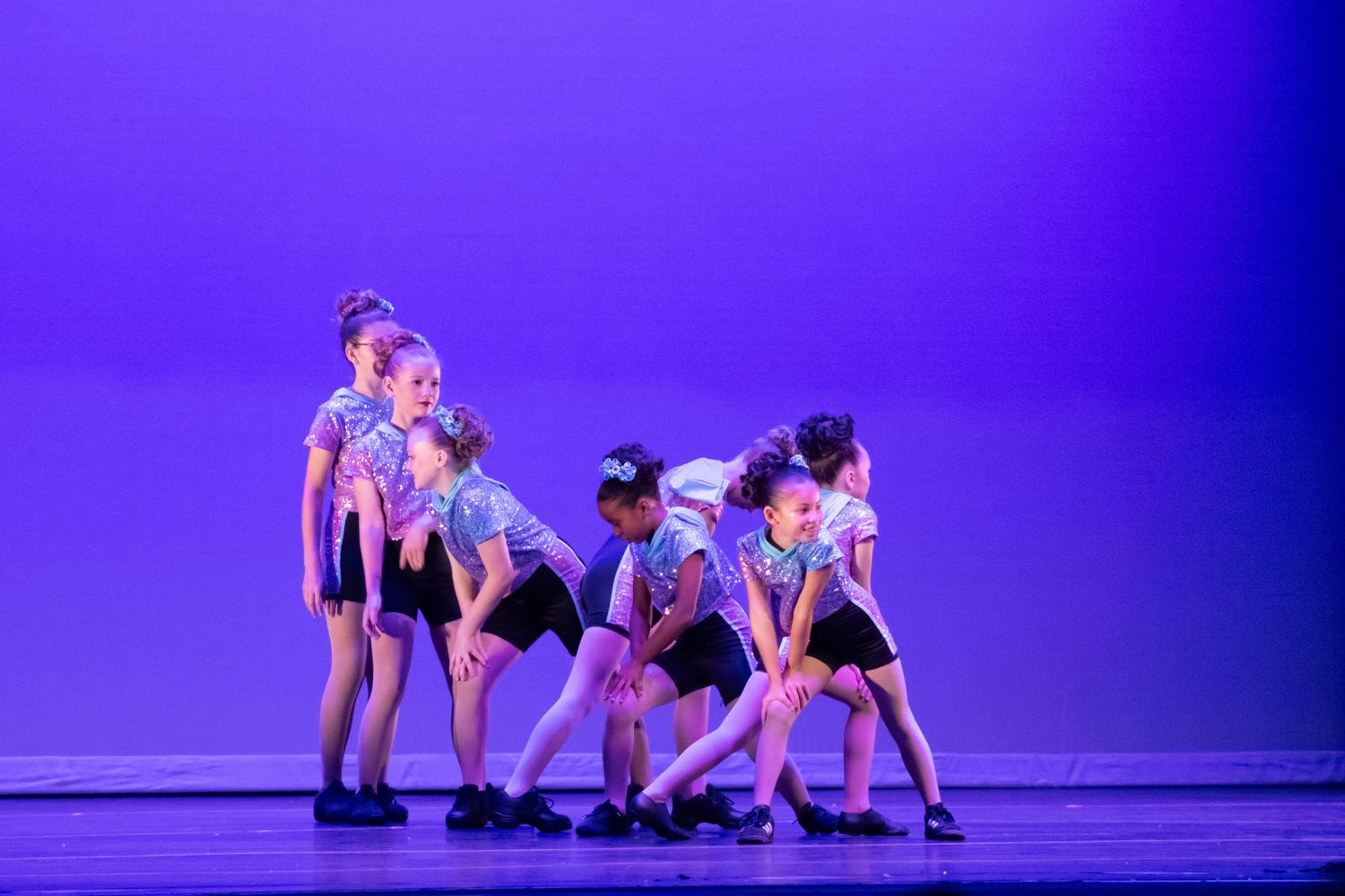 Young dancers in blue tops perform a group routine on a purple-lit stage during a dance recital.