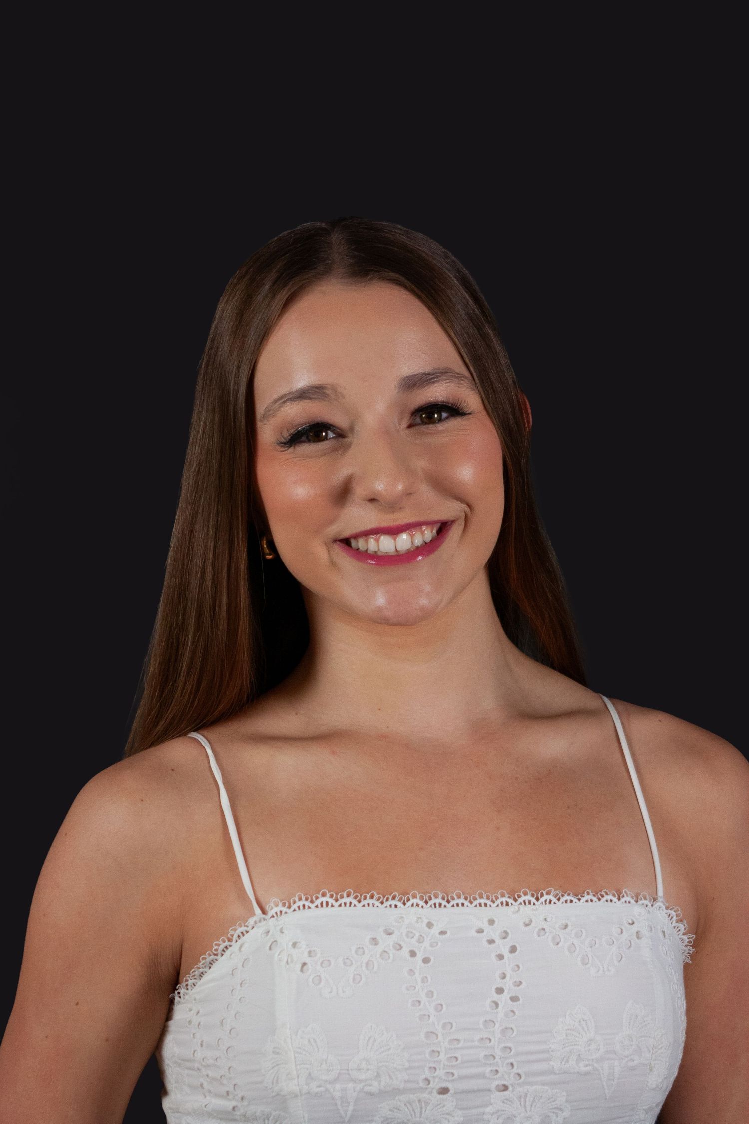 Young woman with long brown hair smiling in white lace top on dark background.
