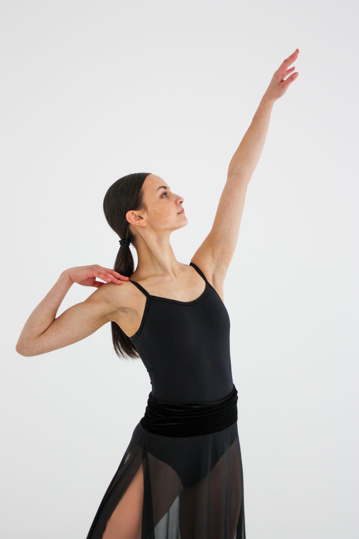 An elegant ballet dancer in a black leotard and skirt raises one arm overhead in a white studio.