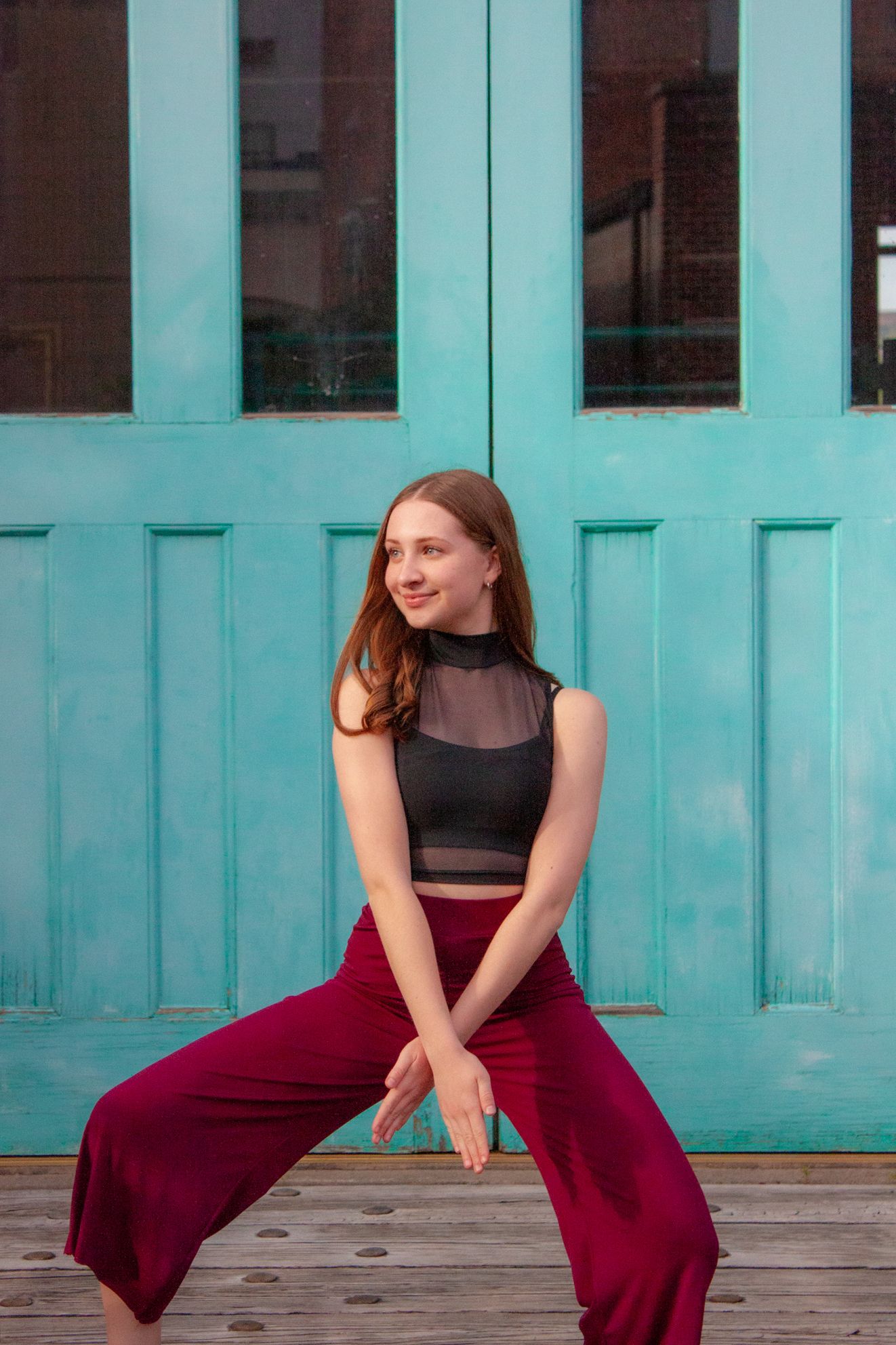 Young dancer in black mesh top and red wide-leg pants poses before a turquoise door.