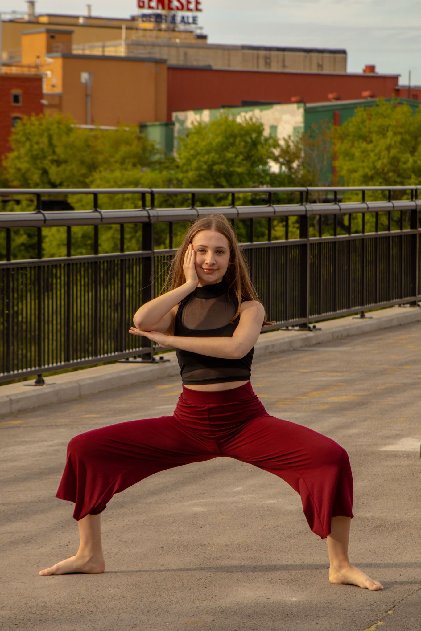 A barefoot dancer in a black top and red wide-leg pants performs a wide stance pose on a rooftop.