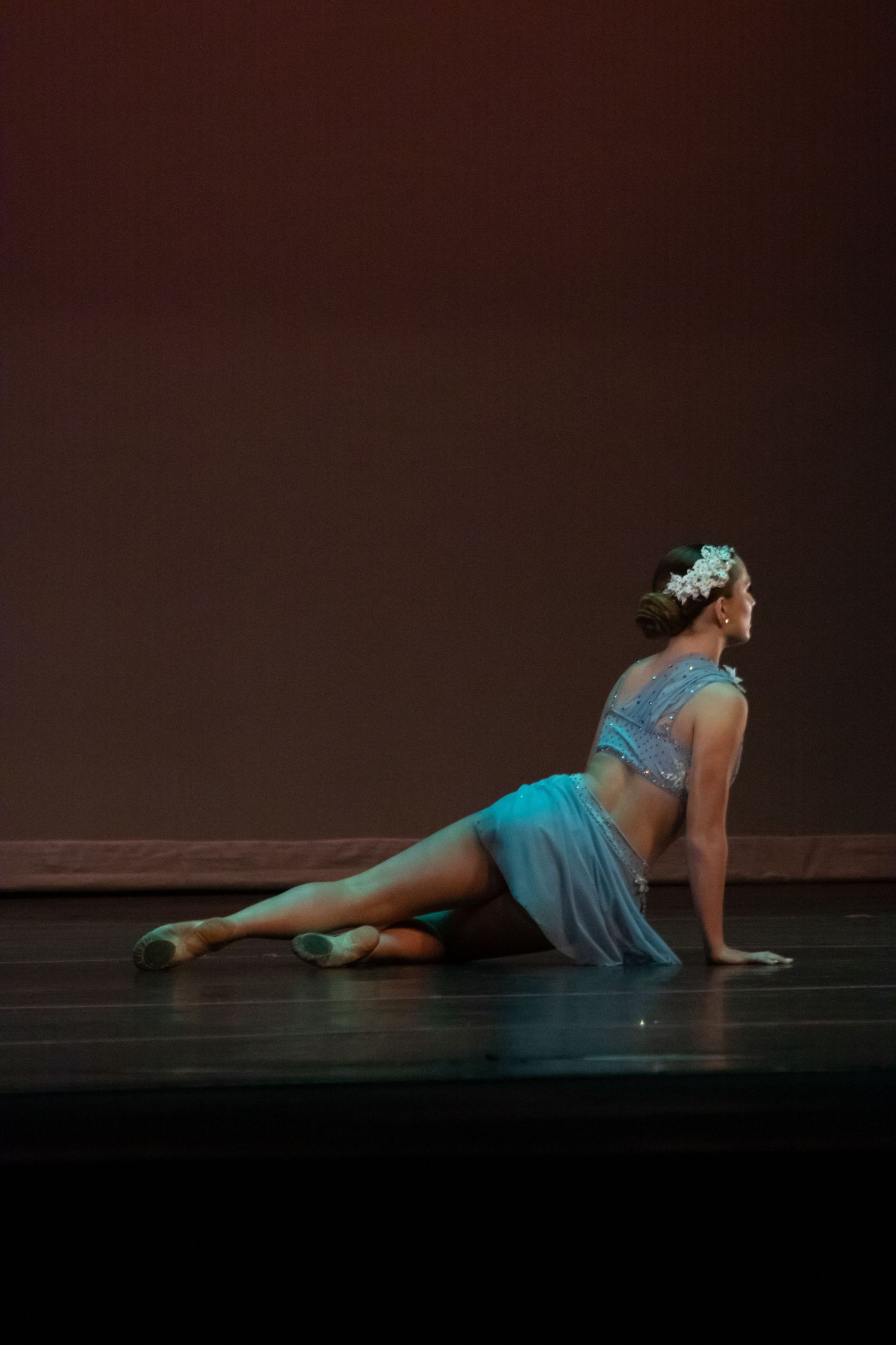 A dancer in a turquoise costume with floral headpiece performs a graceful seated pose on stage.