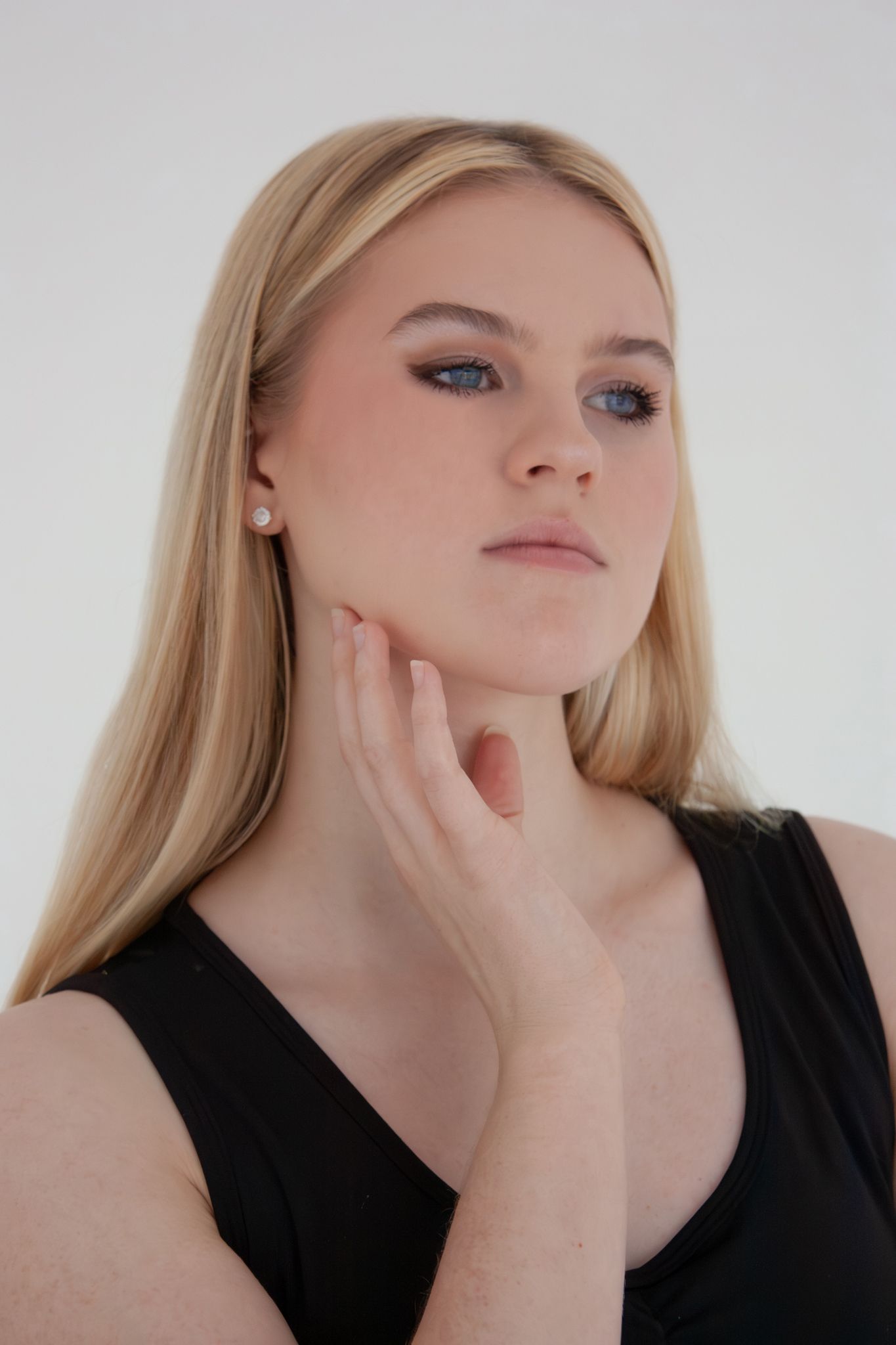 Blonde woman in black tank top touching her chin against white background.