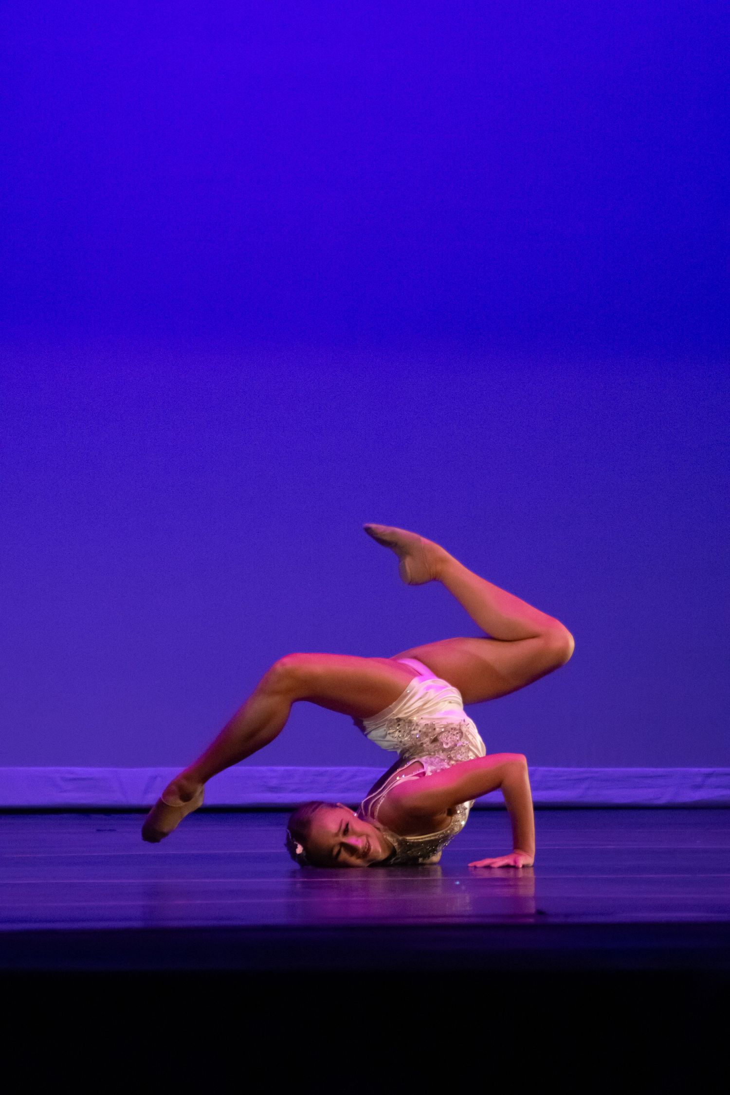 A flexible dancer performs an advanced backbend floor pose with legs raised on a blue-lit stage.