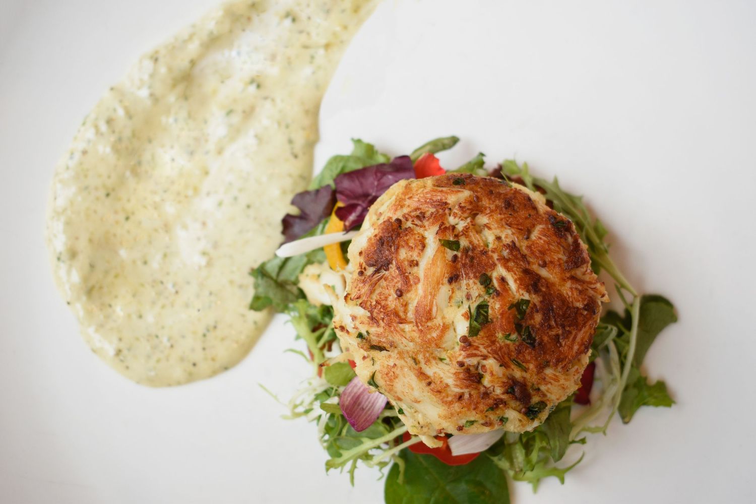 Pan-seared crab cake over fresh mixed greens and vegetables with creamy herb aioli on white plate.