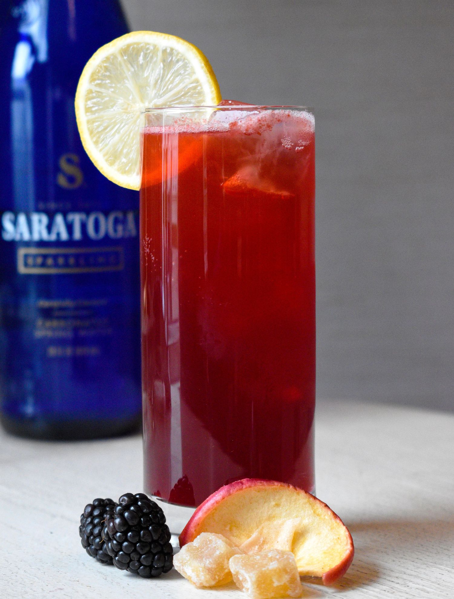Tall red berry cocktail garnished with a lemon wheel, blackberries, and peach slices with Saratoga water in background.