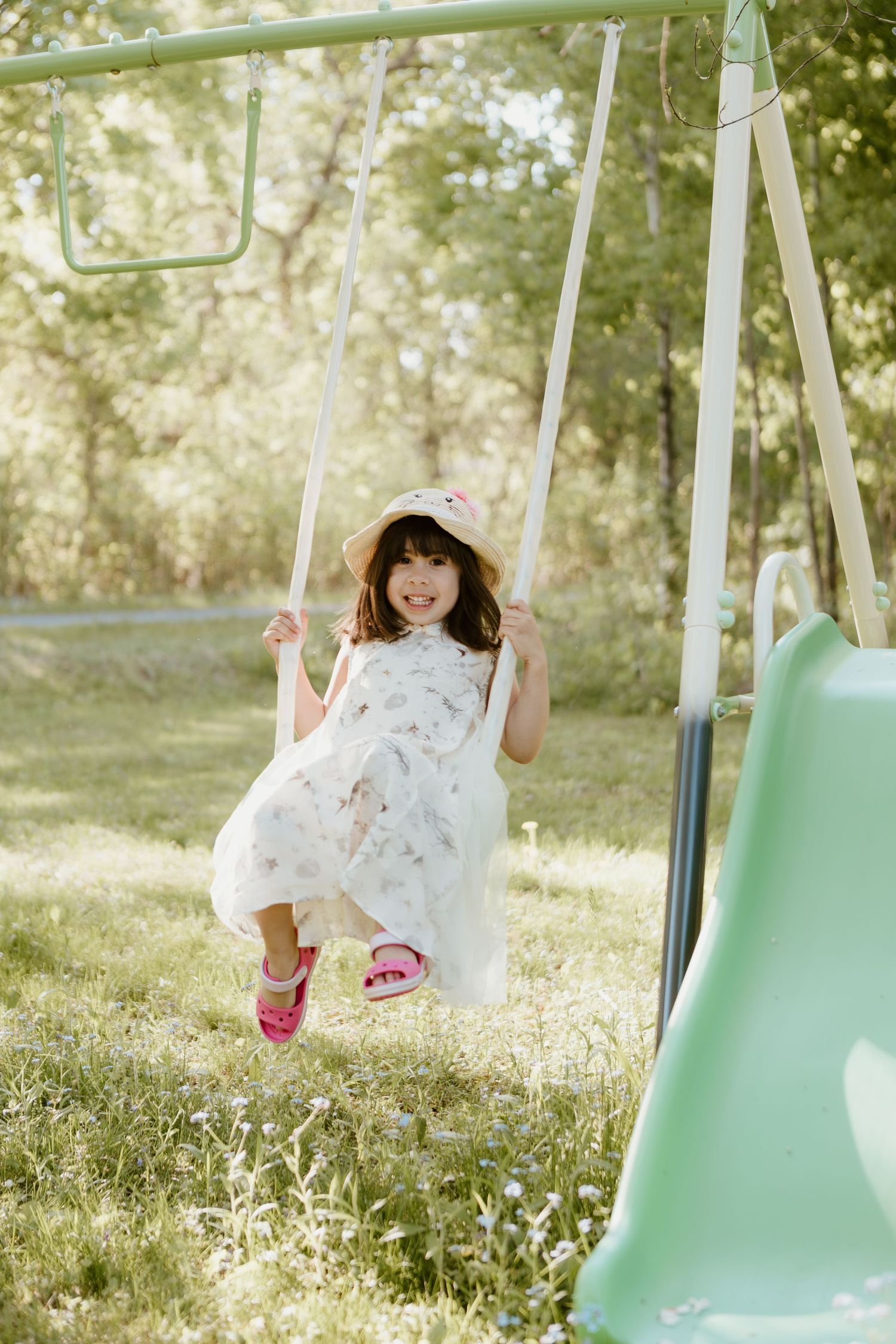 A young girl in a white floral dress and sun hat smiles while swinging on a backyard swing set on a sunny day.