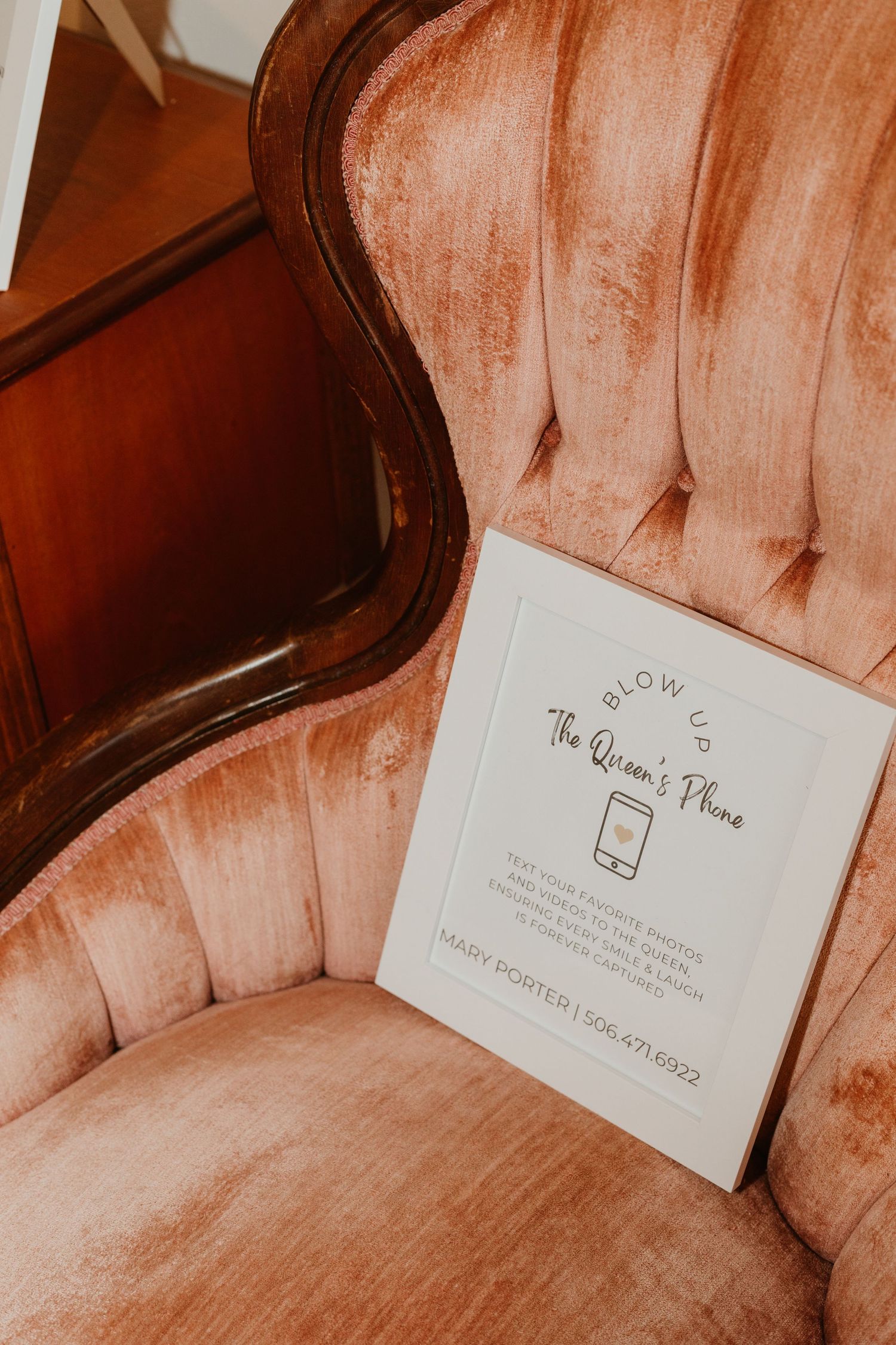 A white sign on a pink velvet tufted chair reading Blow Up The Queens Phone with contact details.
