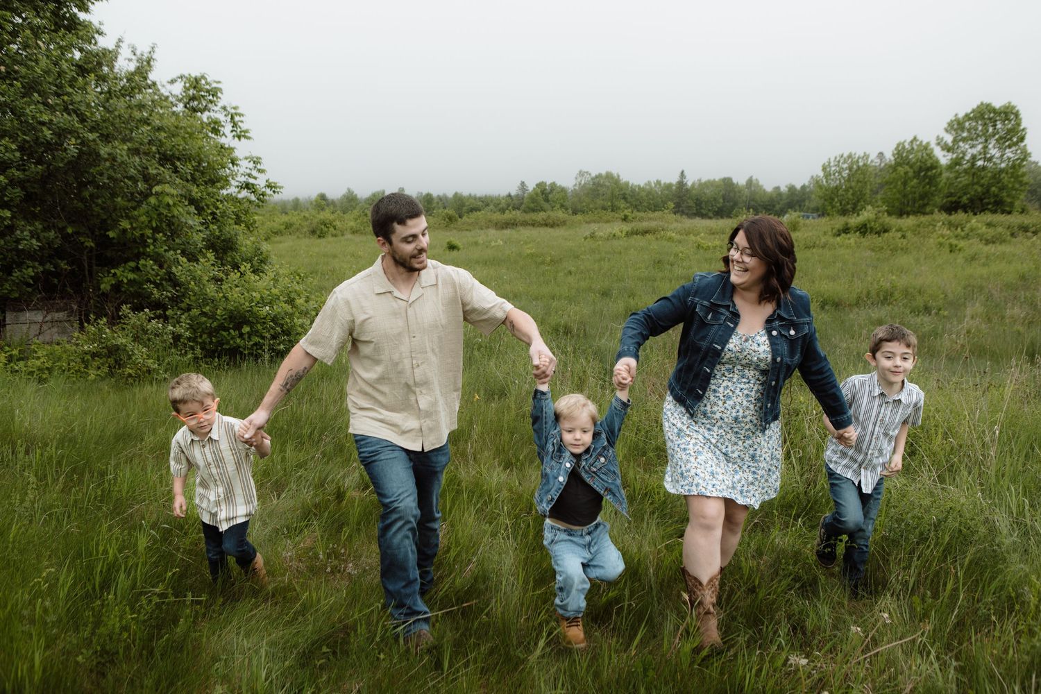 Family walking through a lush green meadow, parents holding hands with three young boys on a cloudy day. New Brunswick Family Photography