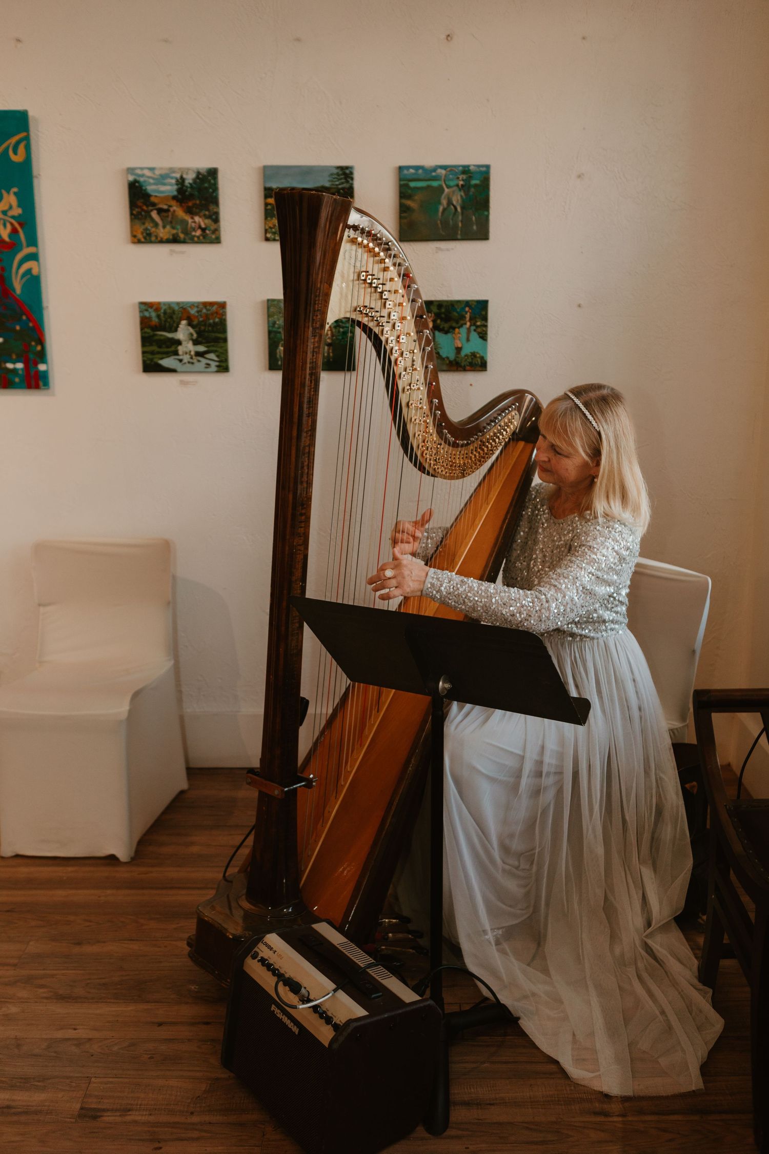 Blonde harpist in white gown playing a concert harp at an indoor venue with colorful artwork on walls. The Gallery on Queen, Fredericton NB