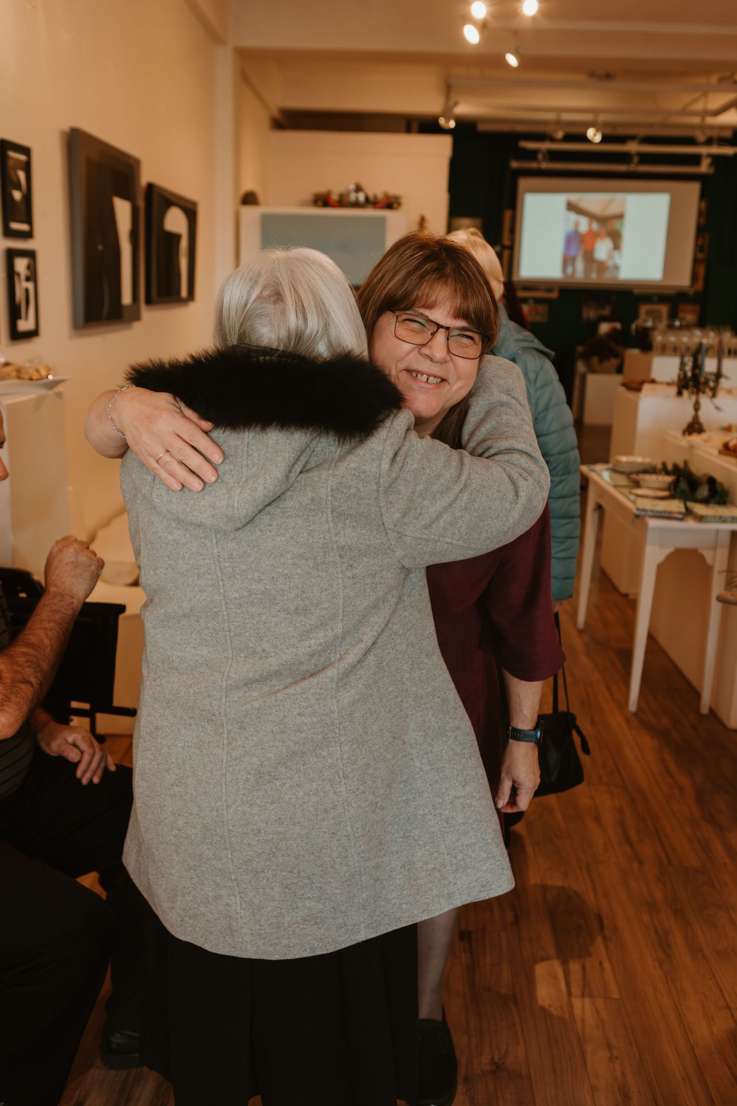 Two women share a warm hug at an art gallery event, smiling joyfully in a cozy indoor setting.