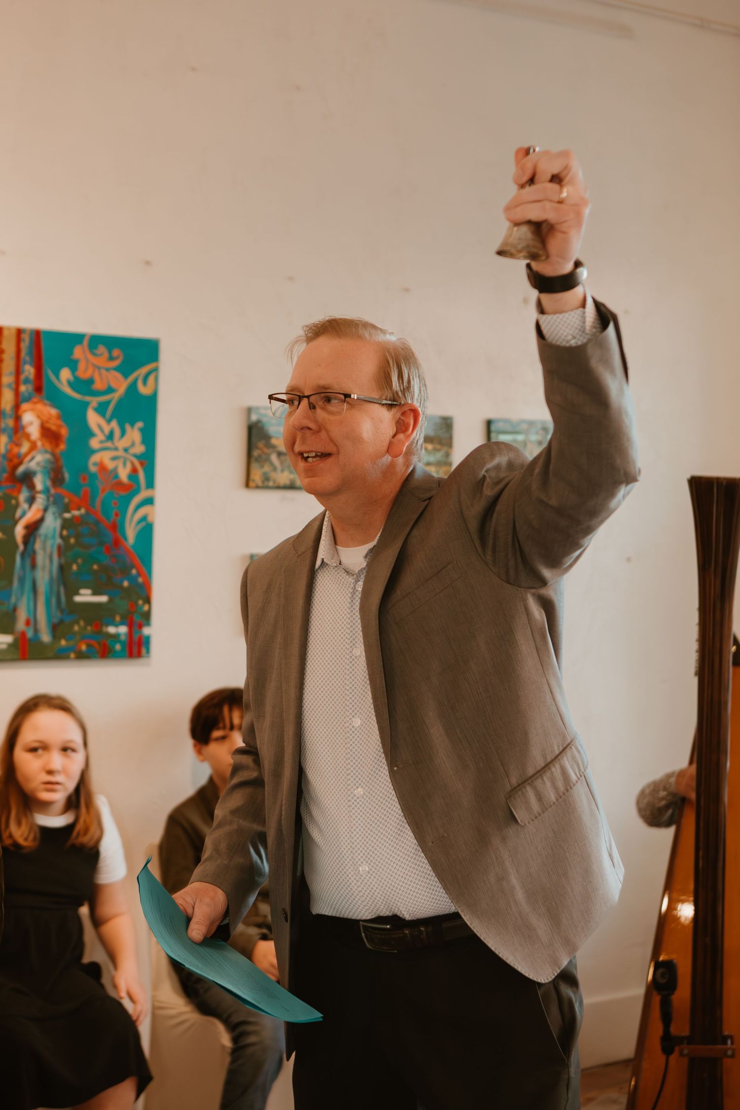Man in gray blazer raises bell at art gallery event with colorful paintings on white walls in background.