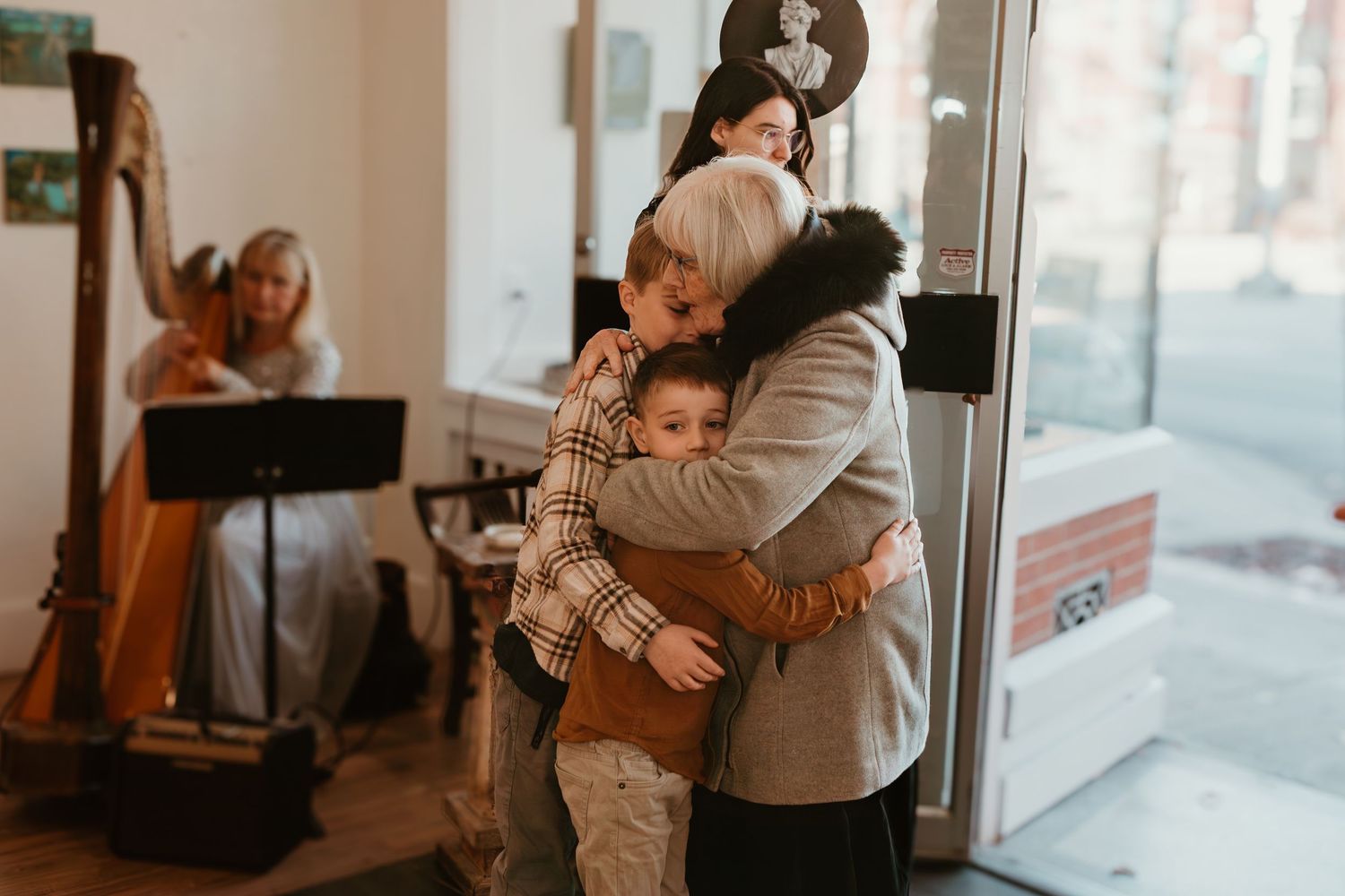 Family group hug near a window with a harpist playing in the background at a warm indoor gathering.