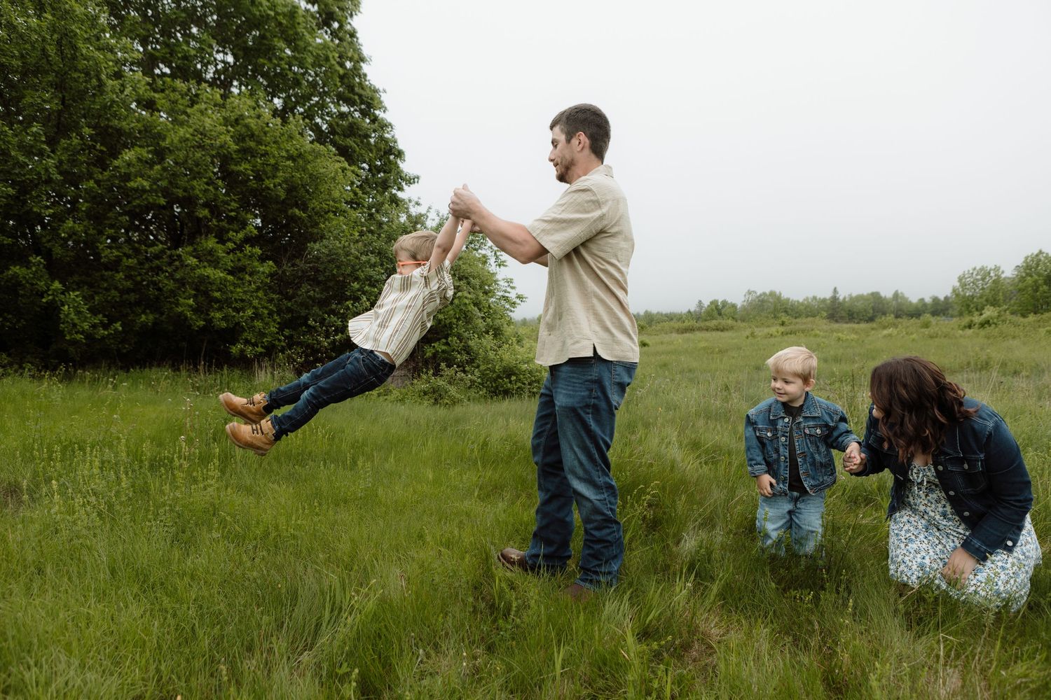Family playing in a green meadow, father swinging child in air while mother watches with toddler nearby. New Brunswick Family Photography