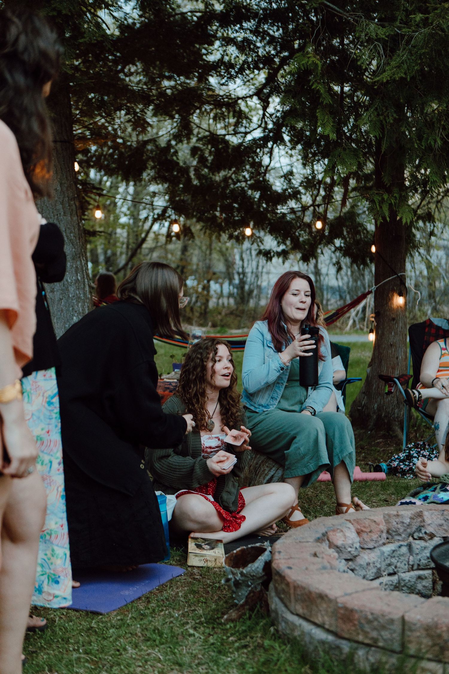 Women gather around a fire pit at an outdoor evening party under string lights and trees.