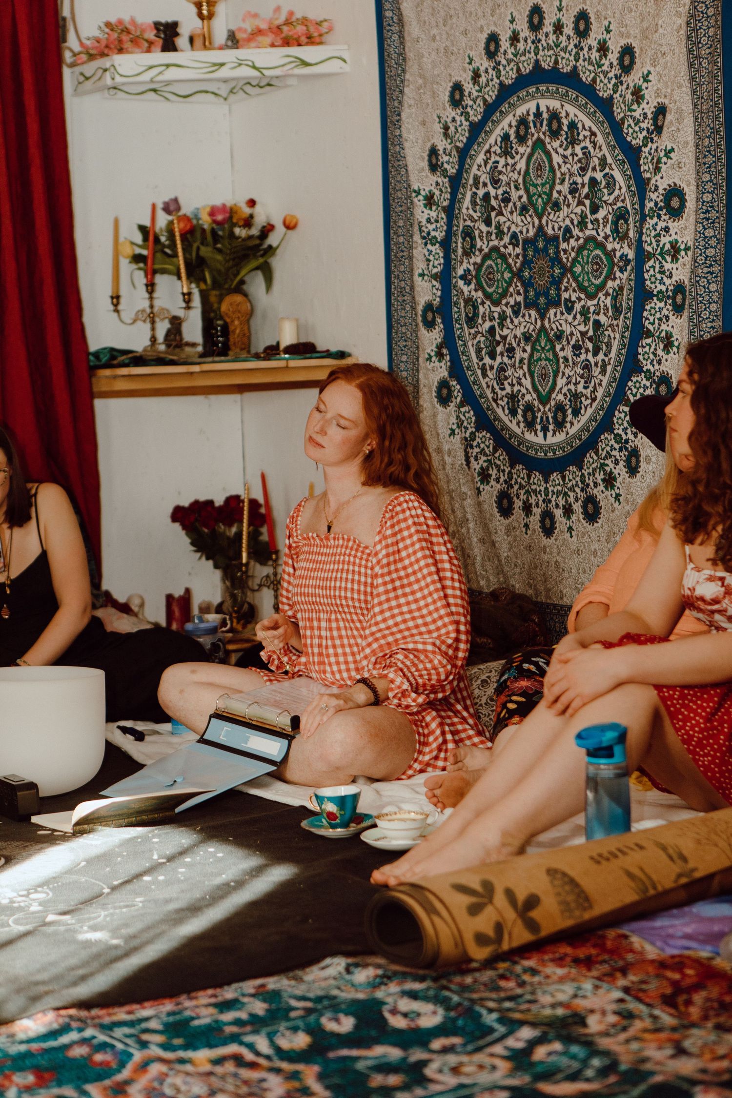Women sitting cross-legged on floor with mandala tapestry, candles, and flowers in a cozy bohemian meditation space.