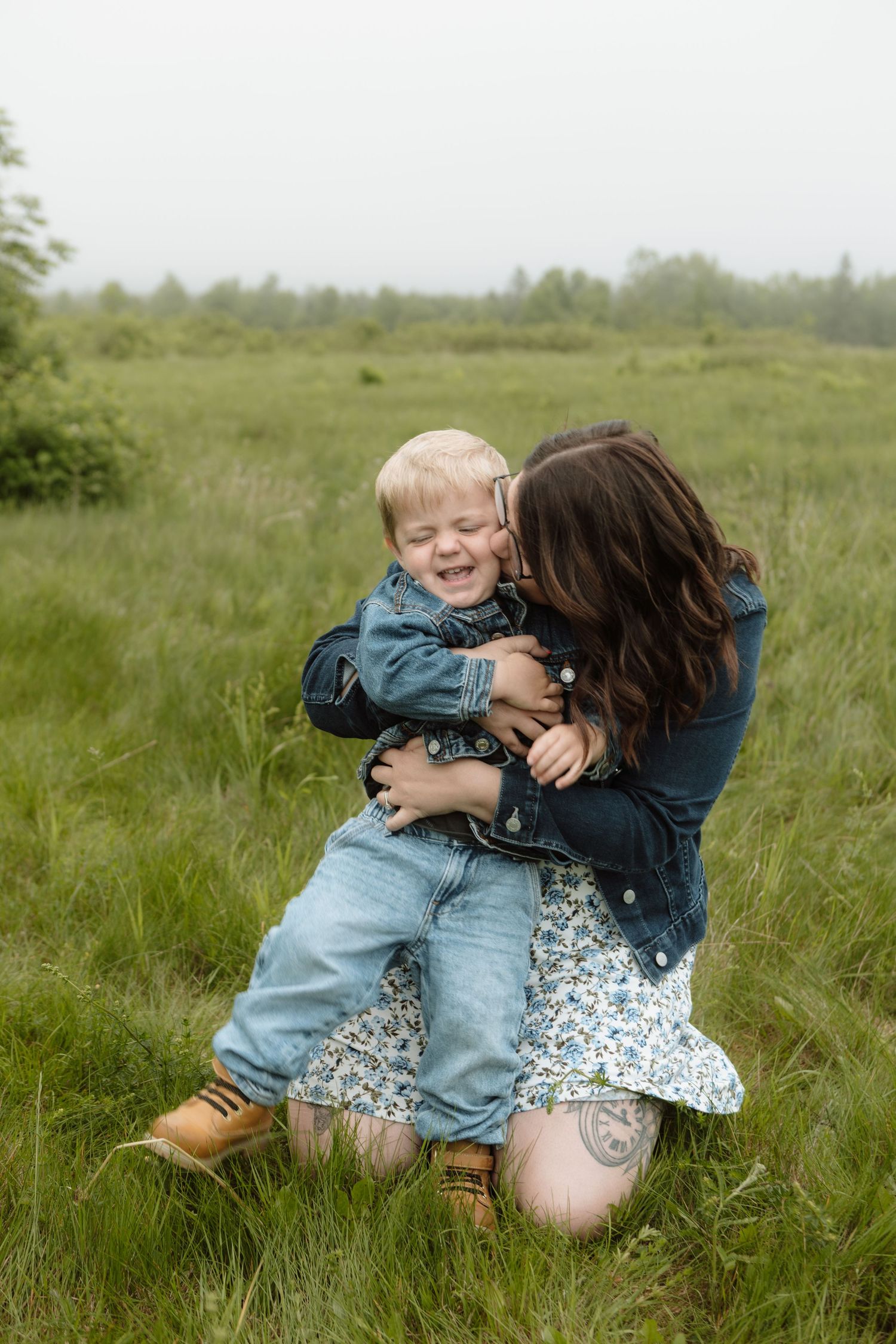A mother in a green field, hugging and kissing her laughing toddler son, both wearing denim outfits. New Brunswick Family Photography
