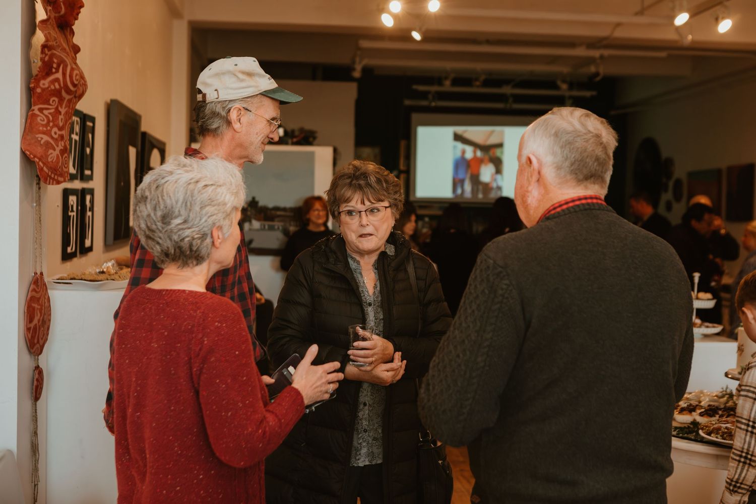 Four people engaged in lively conversation at an art gallery opening, surrounded by artwork on display.