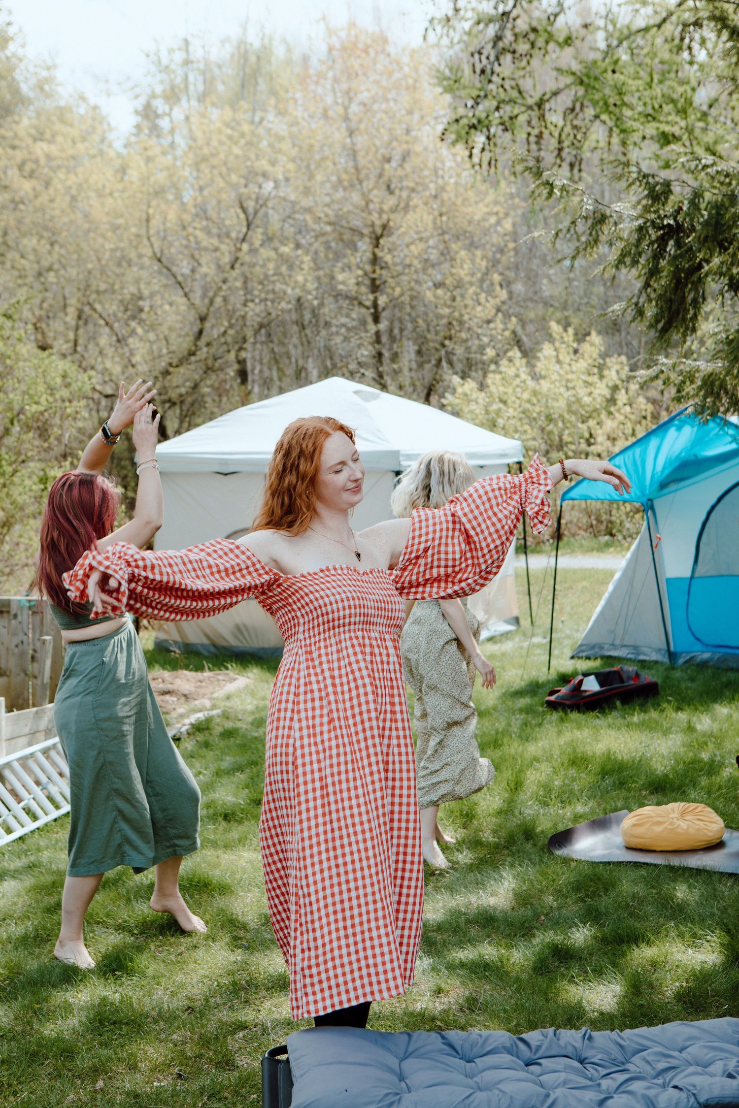 Two women dancing outdoors at a campsite surrounded by tents and trees on a sunny day.