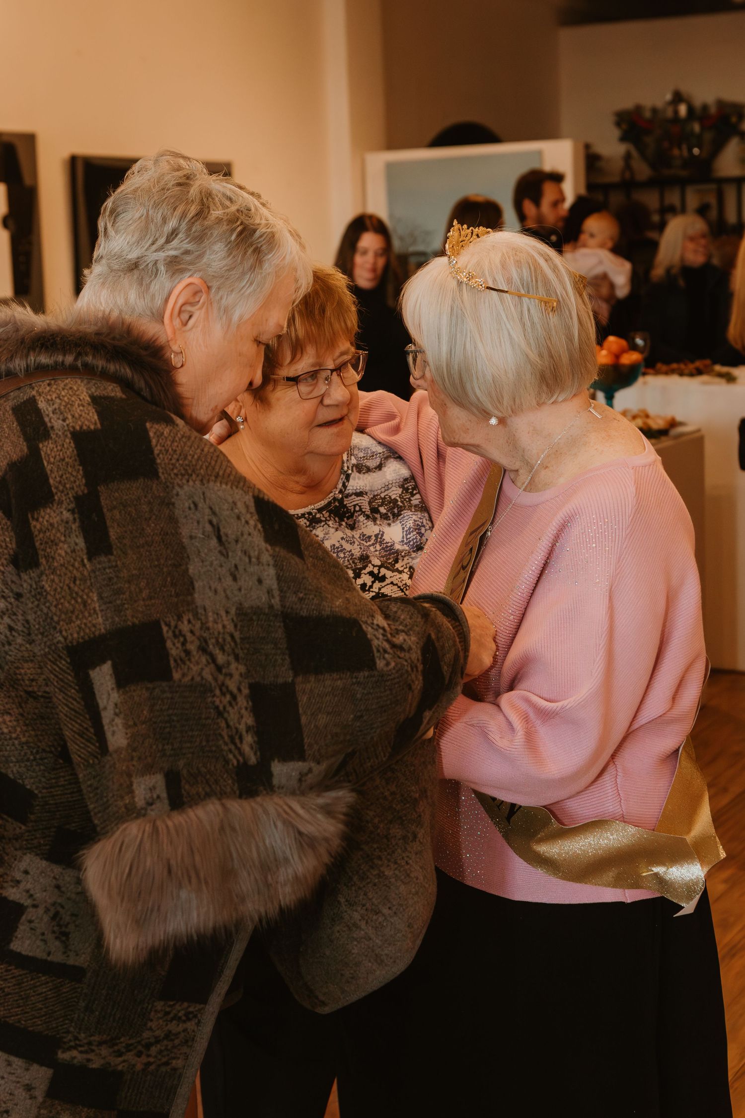 Three elderly women sharing a warm embrace at an indoor social gathering with guests in the background.