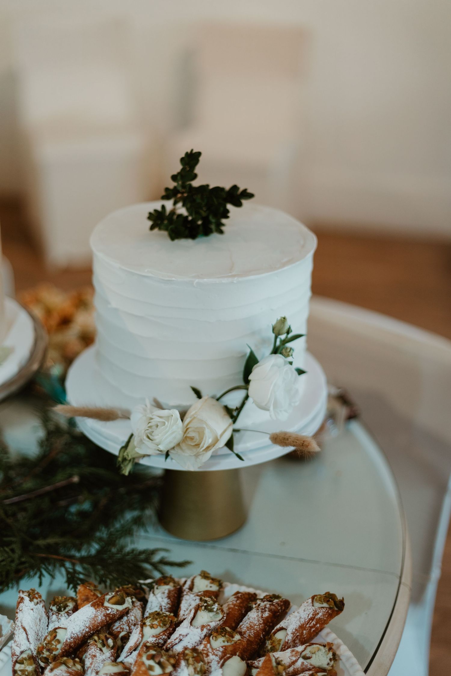 Single-tier white wedding cake with roses and greenery on gold stand, surrounded by cannoli on glass table.