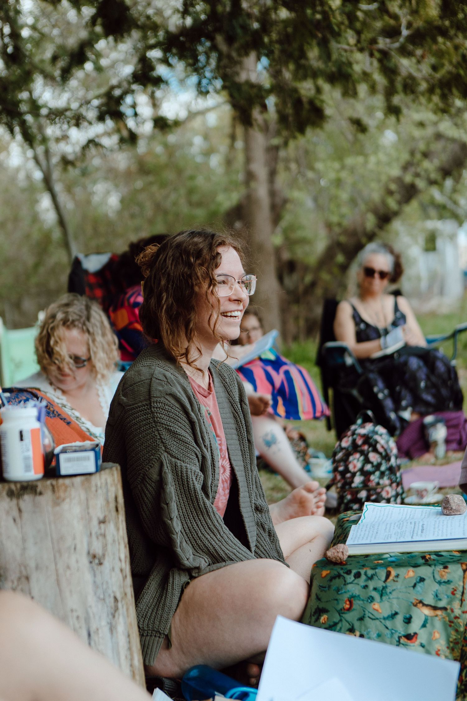 A smiling woman with glasses sits outdoors at a casual gathering among trees, wearing a green cardigan.