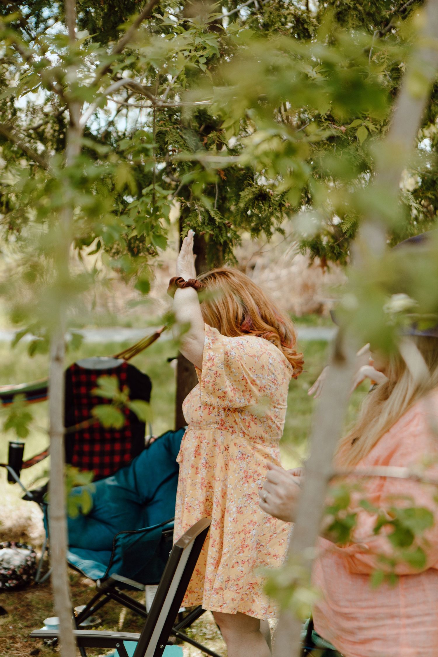 Woman in floral dress reaching up to touch tree branches at an outdoor garden gathering with chairs visible.