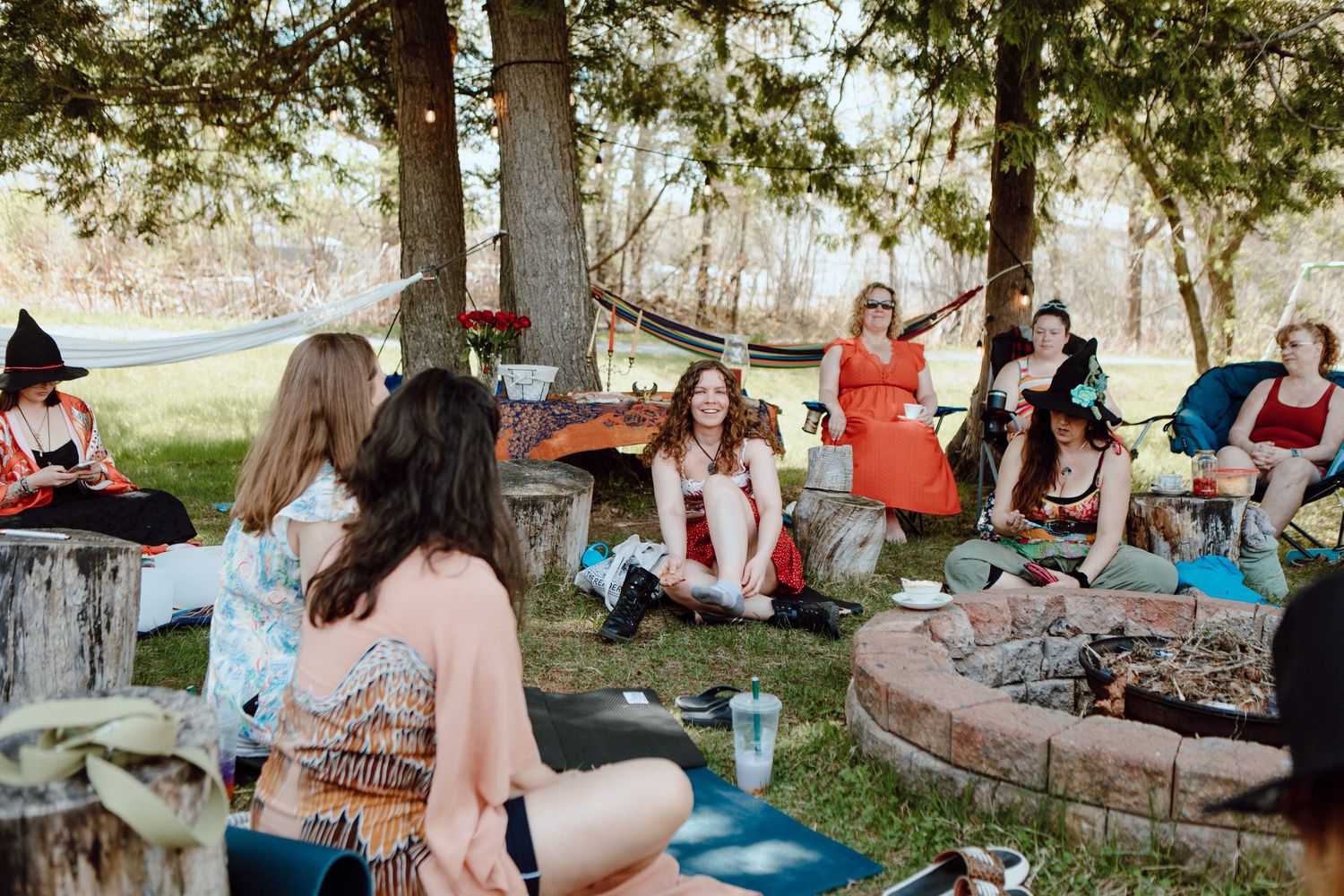 Women gathered around a rustic fire pit outdoors, enjoying a festive Halloween-themed garden party under trees.